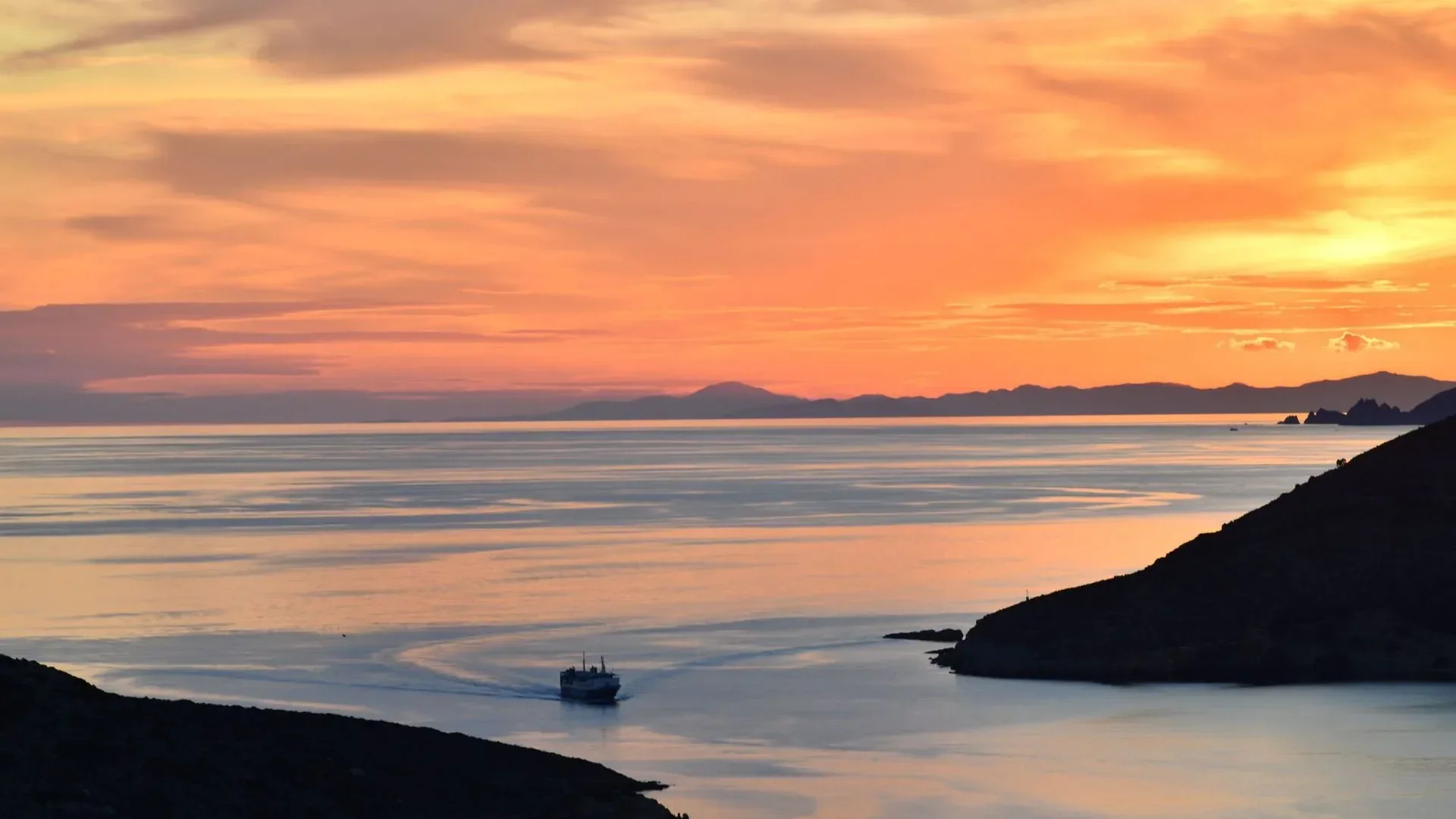 A small ferry crossing calm water at sunset, dark headlands on either side and distant hills under an orange sky.