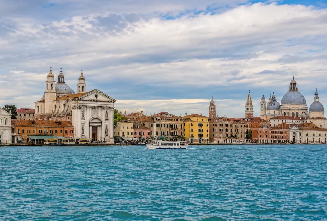 A quiet Giudecca waterfront in late afternoon, moored boats and pale buildings, the city visible across the lagoon.