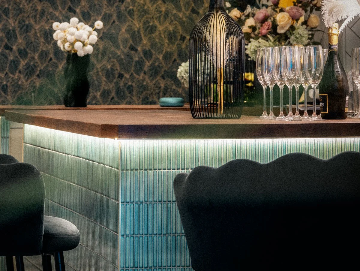 Interior of a bar or lounge area with a wooden counter, glassware, a black decorative lantern, champagne bottle, and floral arrangements in the background, featuring tiled panels with LED lighting.