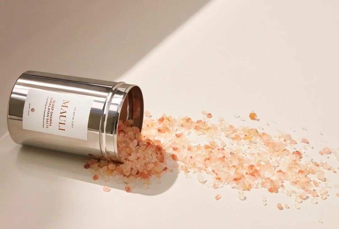 A steel tin of Mauli Rituals Sleep Dharma Himalayan Salts lying open on its side, coarse pink salt spilling across a pale cream surface in soft daylight.