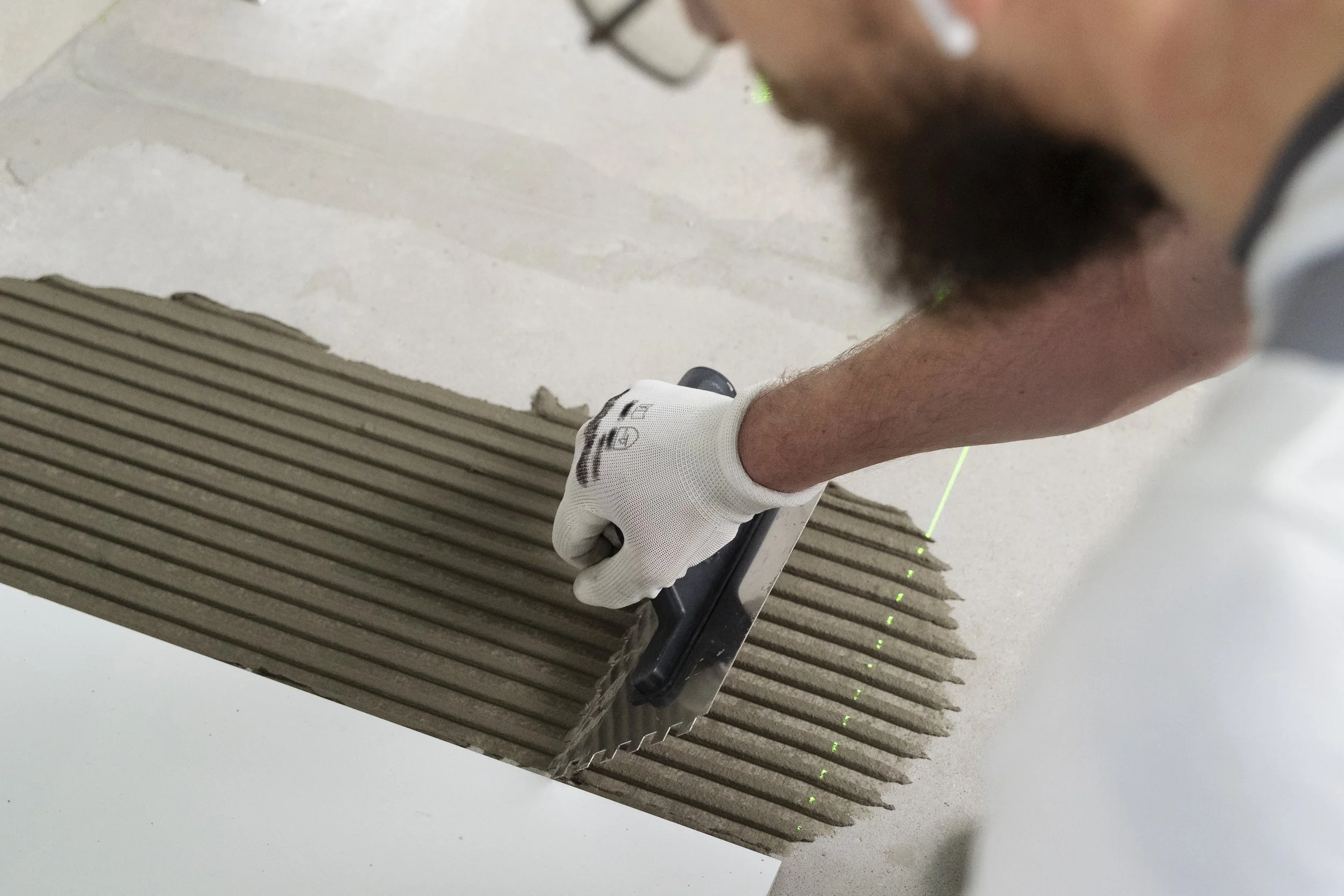 A person wearing white gloves is using a notched trowel to spread adhesive on a large concrete tile with grooves. The person is partially visible, focusing on their arm and hand, and they have facial hair and glasses. There is a green laser level visible on the workspace.