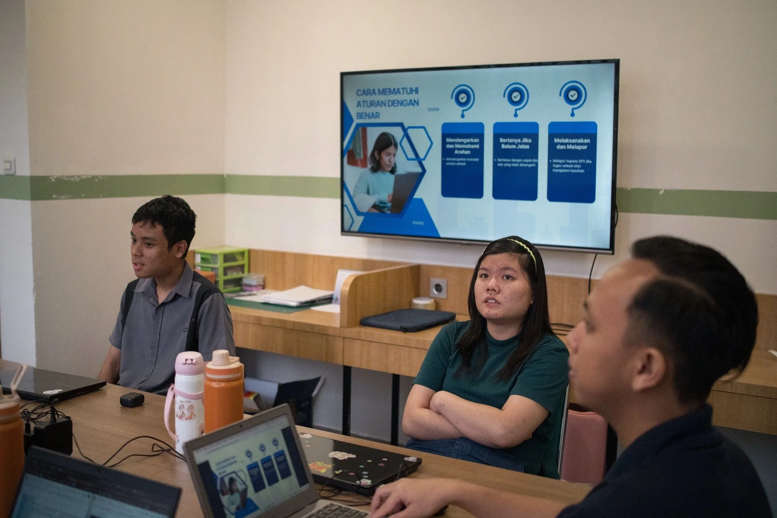 A group of people in a meeting room, sitting at a table with laptops and water bottles, with a large screen displaying a presentation in the background.