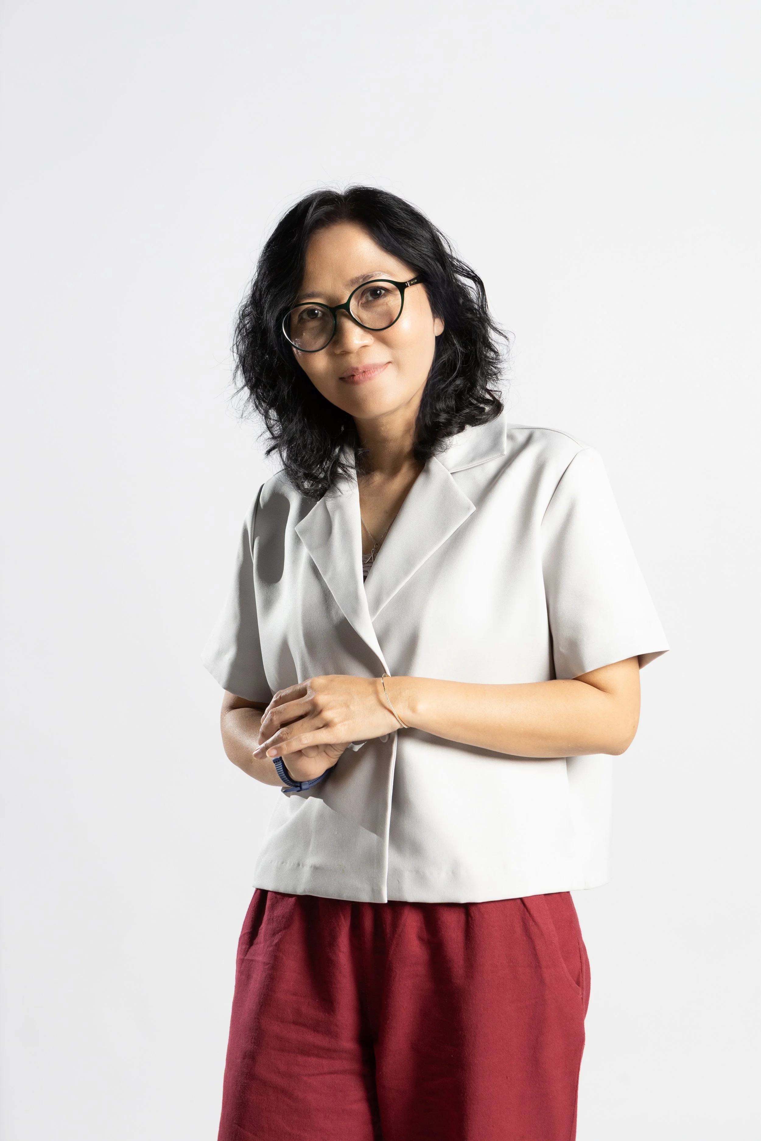 A woman with black, curly hair and glasses wearing a light gray blazer and red pants, standing against a plain white background.