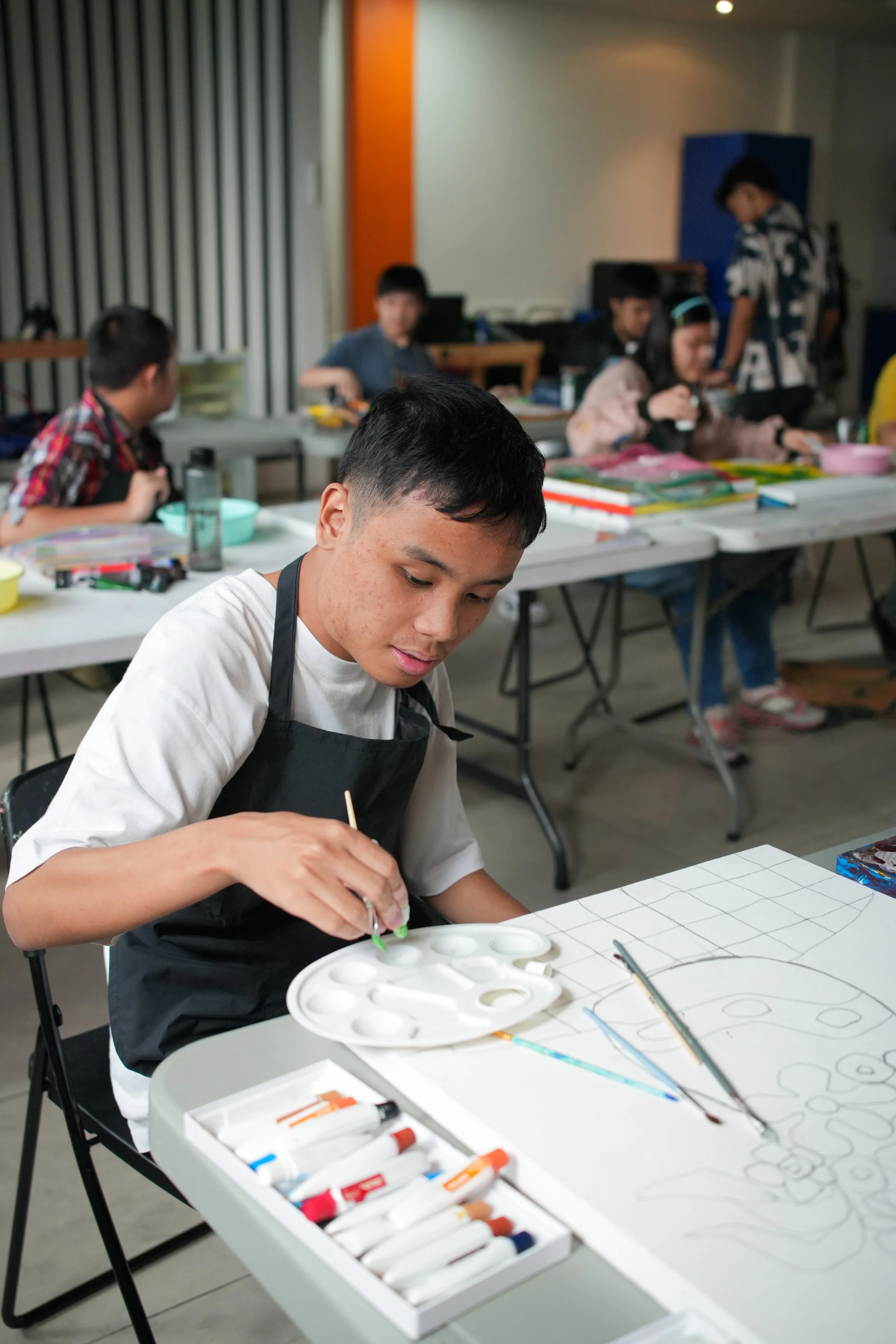 A young man painting with watercolors at a table surrounded by children participating in an art activity.