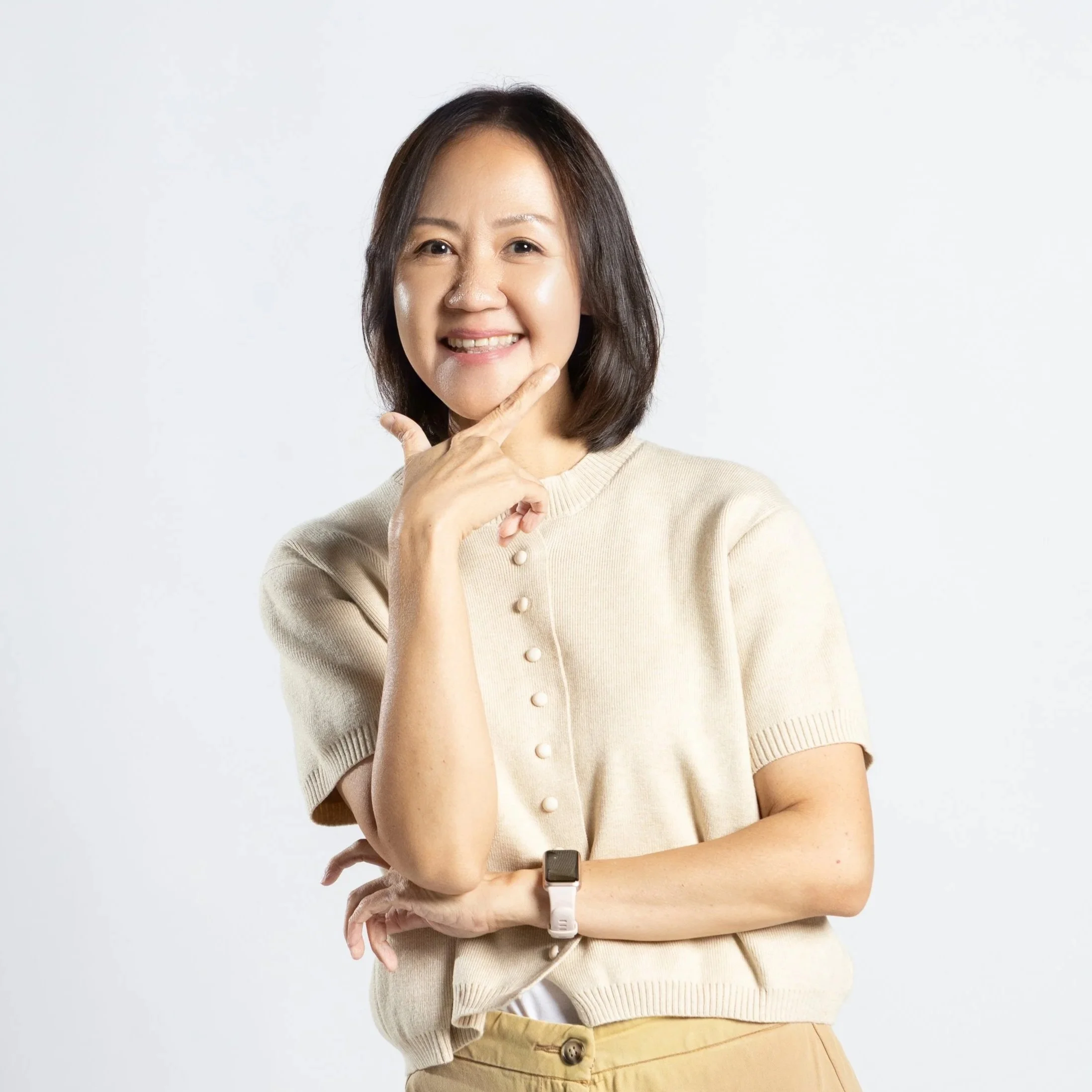 A woman with shoulder-length dark hair, smiling, wearing a beige short-sleeve sweater with pearl buttons, a smartwatch, and beige pants, standing against a white background.