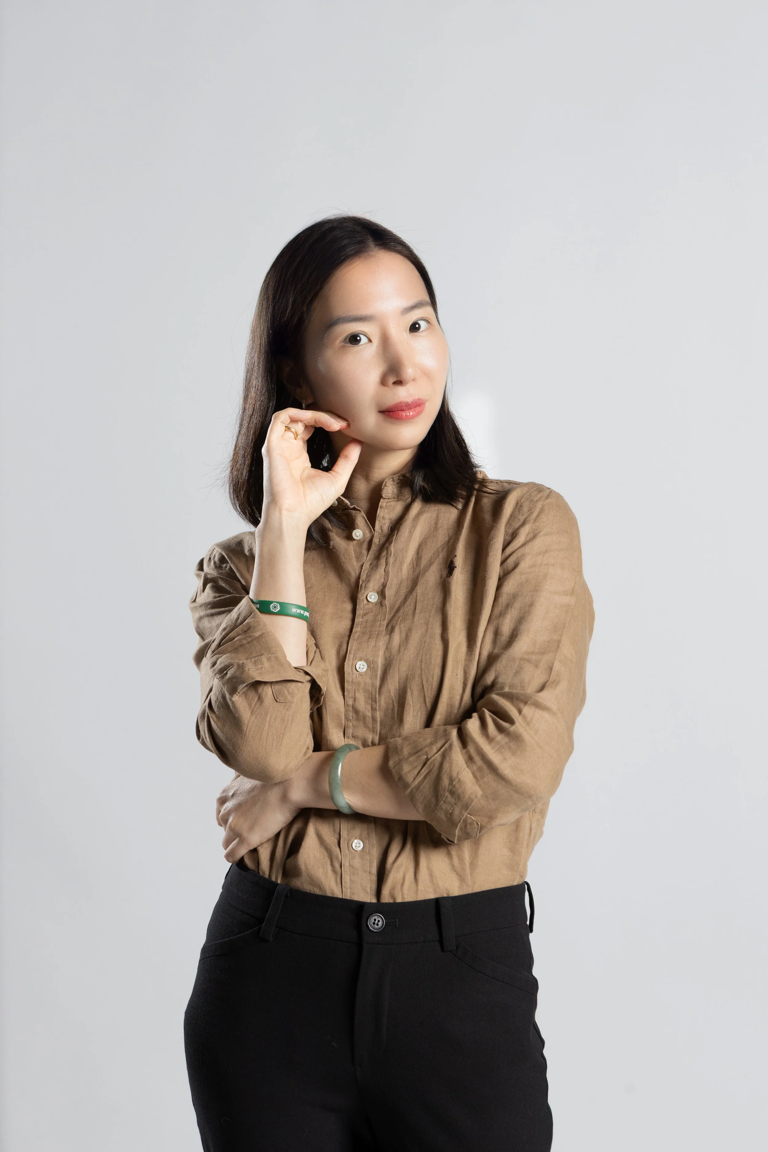 A woman with shoulder-length dark hair, wearing a brown button-up shirt and black pants, standing against a plain white background, looking at the camera with her left hand resting on her chin and her right arm crossed.