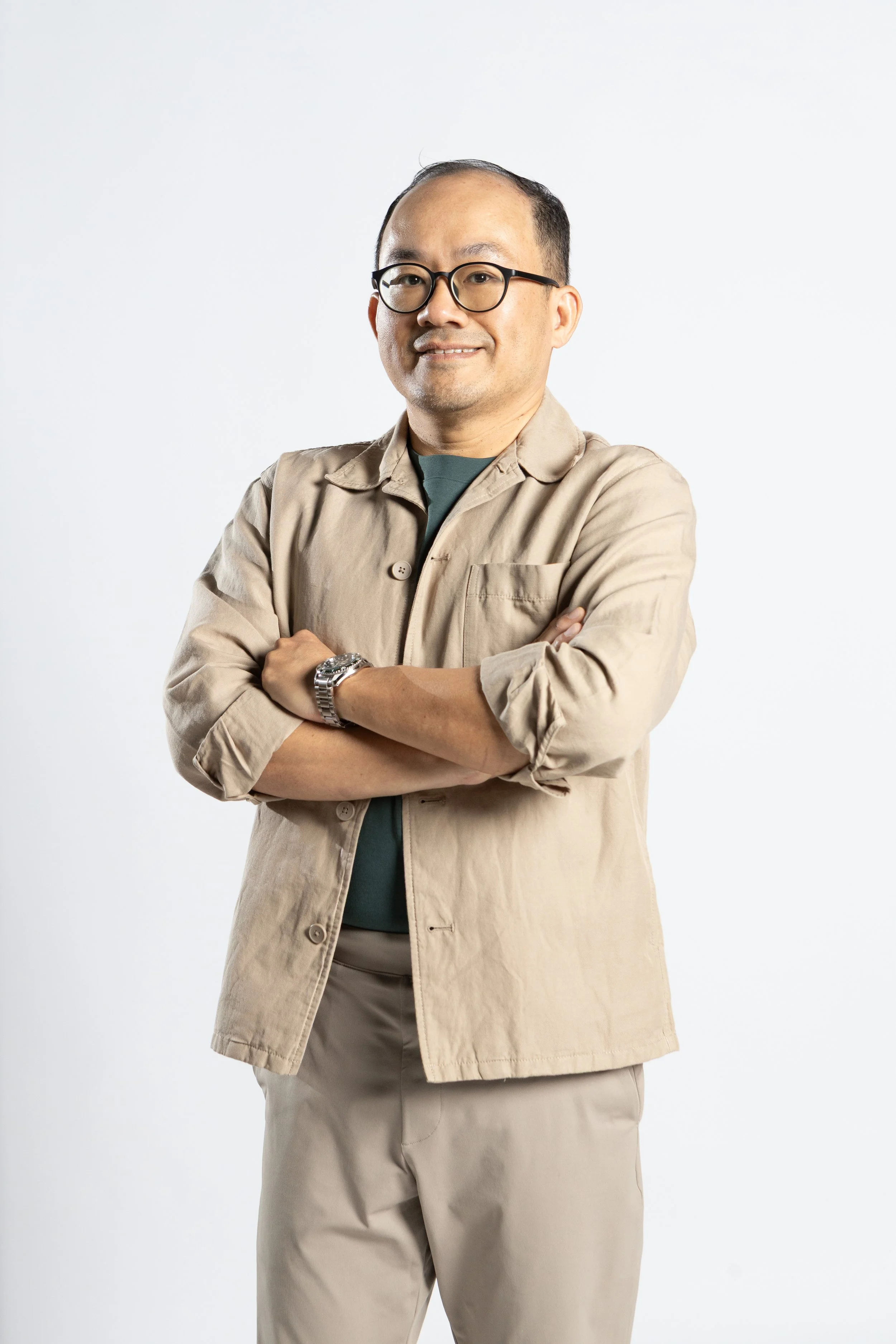 A middle-aged man with glasses, wearing a beige jacket over a dark shirt, standing with arms crossed against a plain white background.