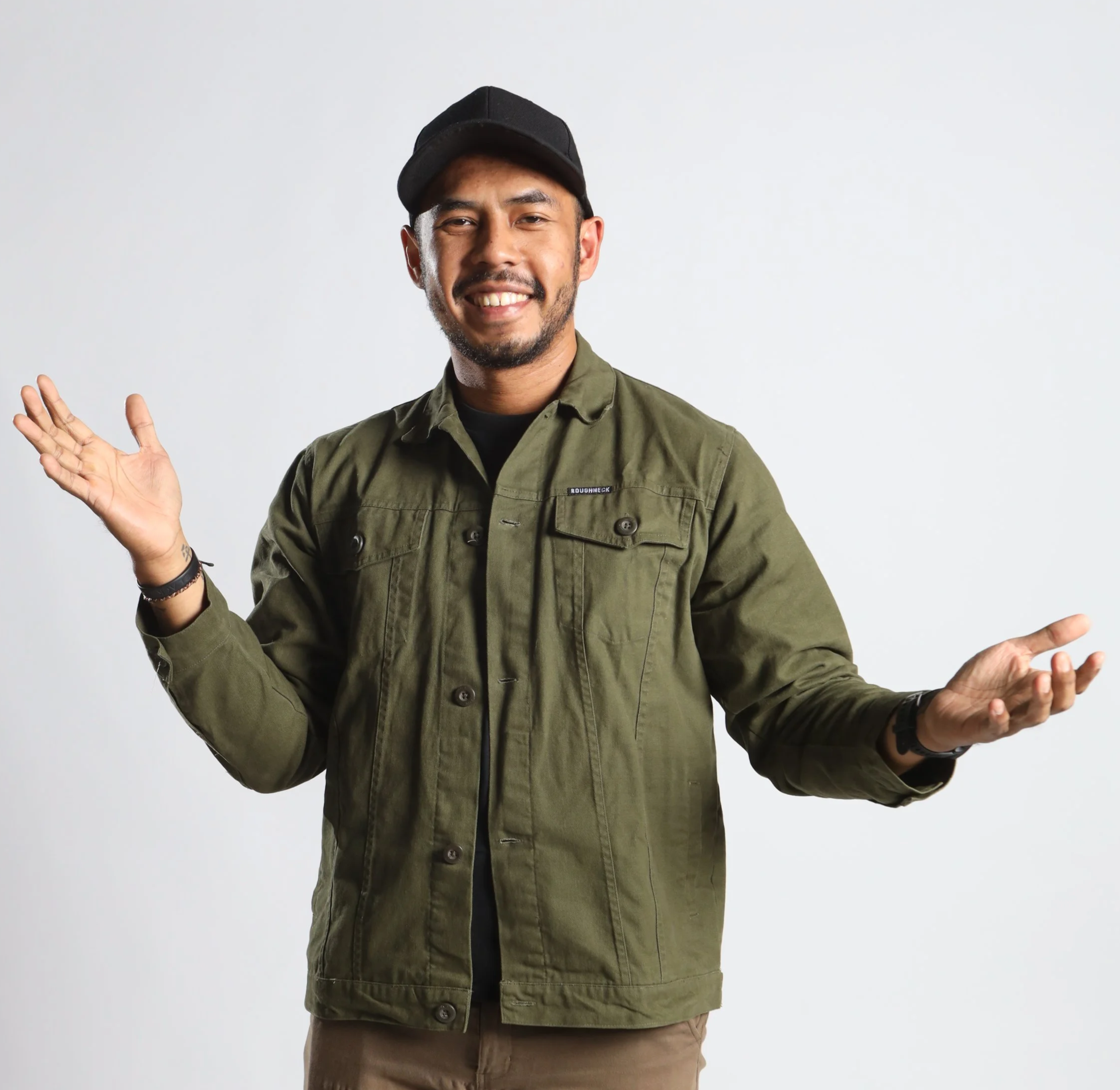 Smiling man wearing a black cap, green jacket, black shirt, and beige pants, gesturing with open arms against a plain white background.