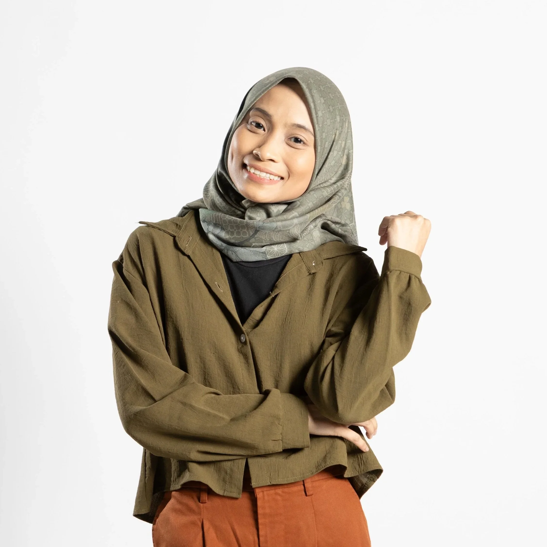 Smiling woman wearing a light patterned hijab, brown jacket, and rust-colored pants, standing against a plain background, with her right arm raised and fist clenched.