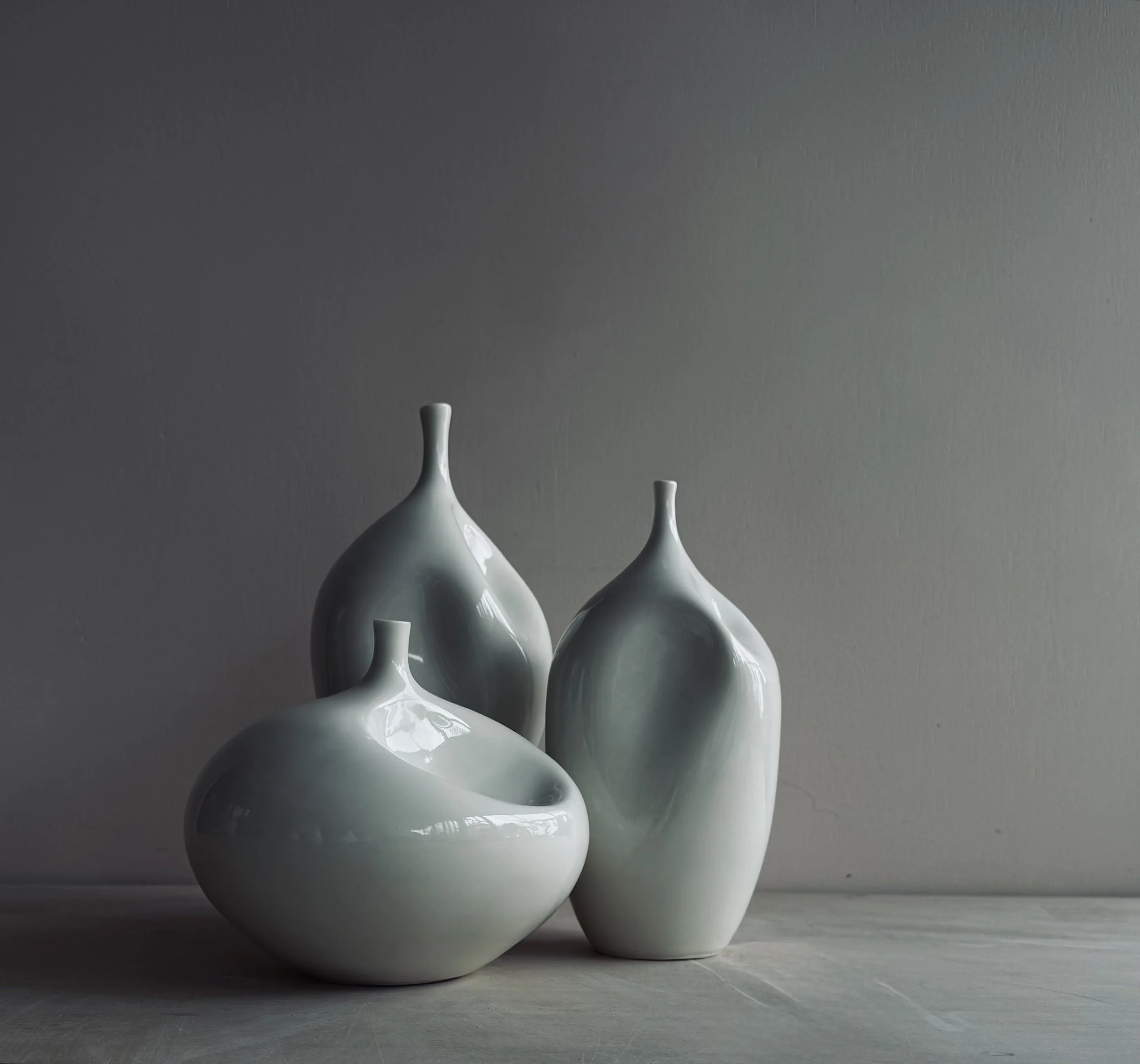 Three white ceramic vases with smooth, rounded shapes and narrow openings, arranged on a light-colored wooden surface against a plain gray wall.