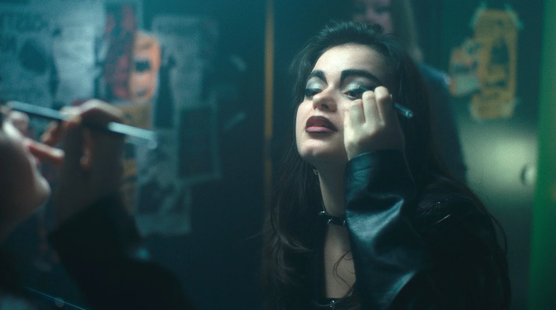A woman with dark hair and makeup sits in front of a mirror, applying makeup with her head tilted back. The setting appears to be a dimly lit room, possibly a makeup studio or bathroom.