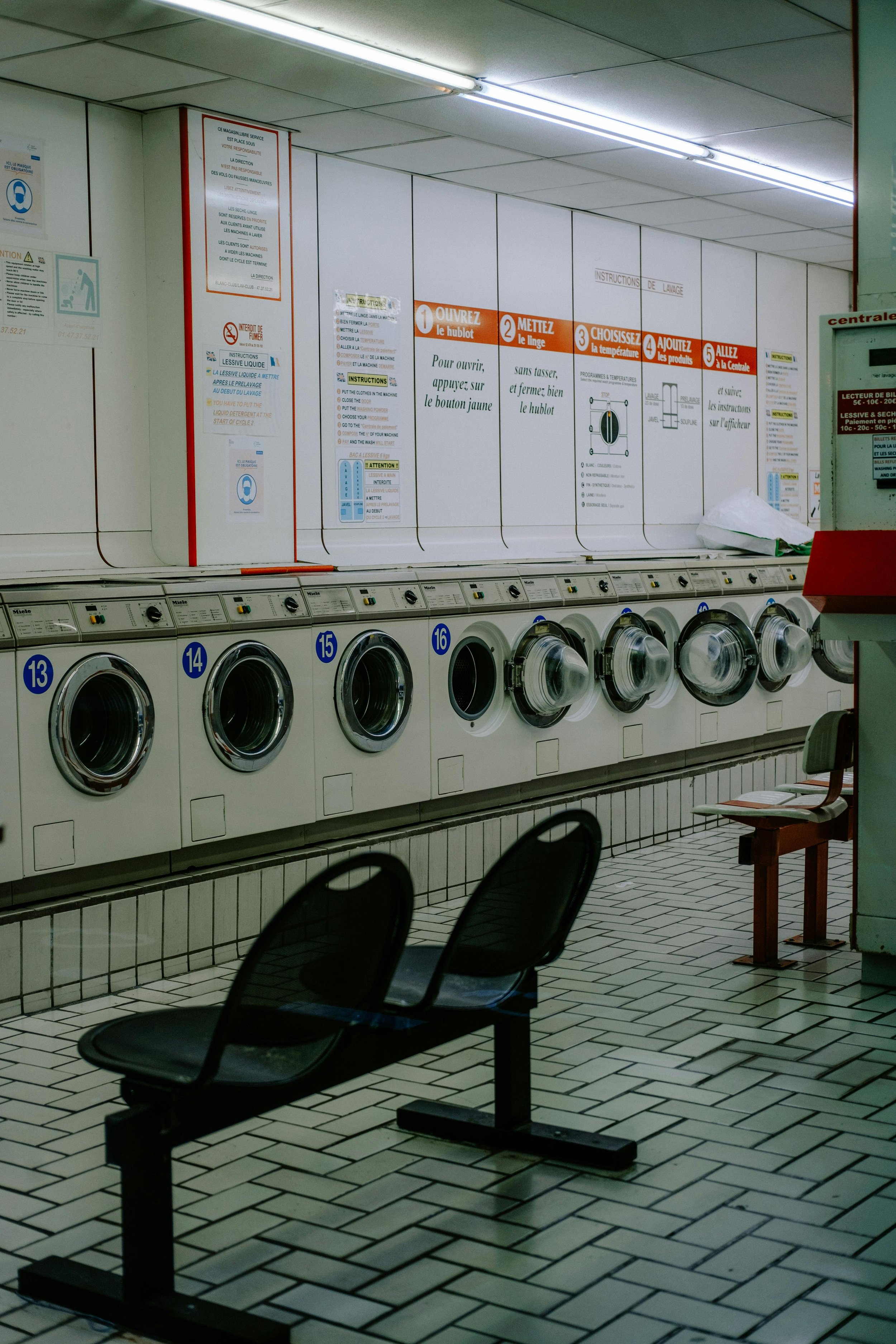 A laundromat with several front-loading washing machines, chairs, and laundry instructions on the wall.