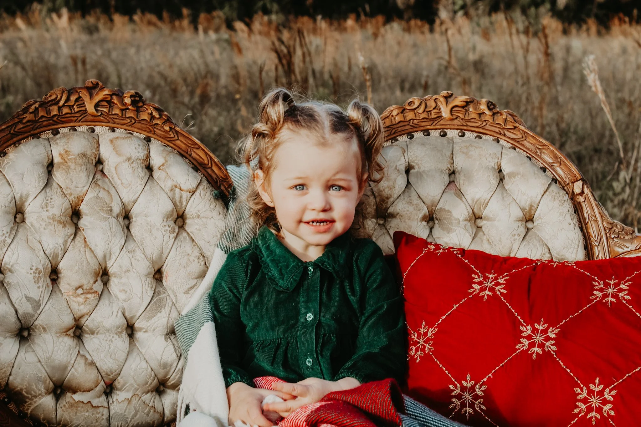 A young girl with curly blonde hair, blue eyes, and a green velvet dress sitting on vintage cream-colored tufted sofa with a red pillow, outdoors in a field.