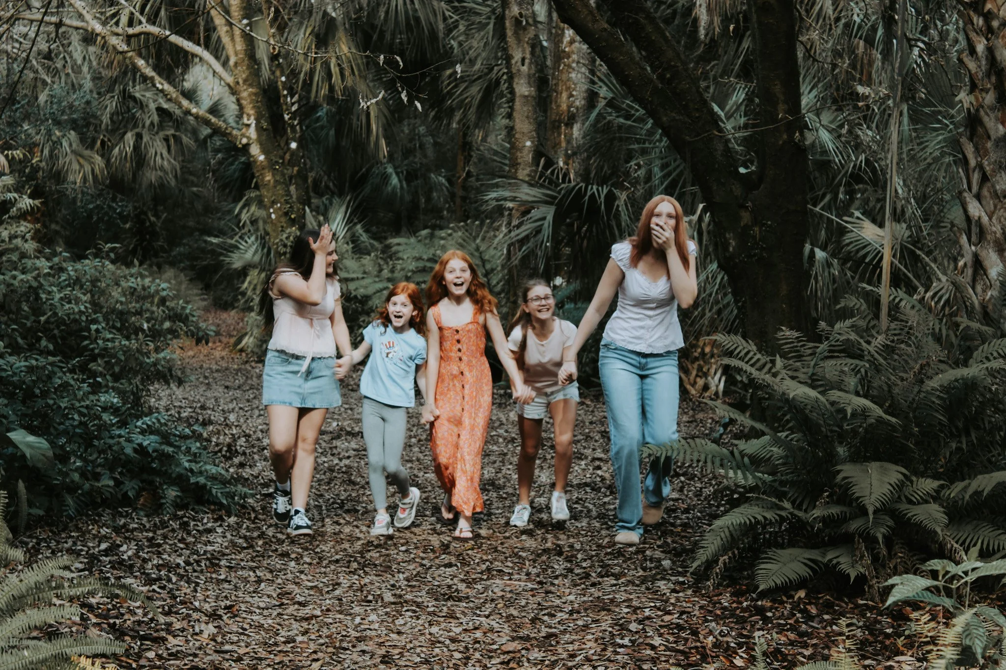 A group of six girls, including a woman, walking and laughing through a forest with lush green plants and trees.