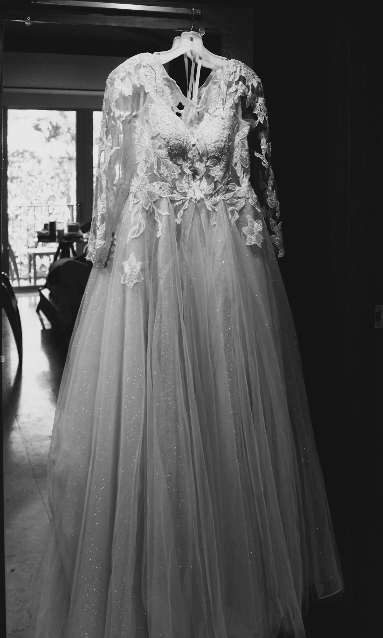 A wedding dress with lace detailing and a full tulle skirt hanging on a hanger in a room with a window in the background.