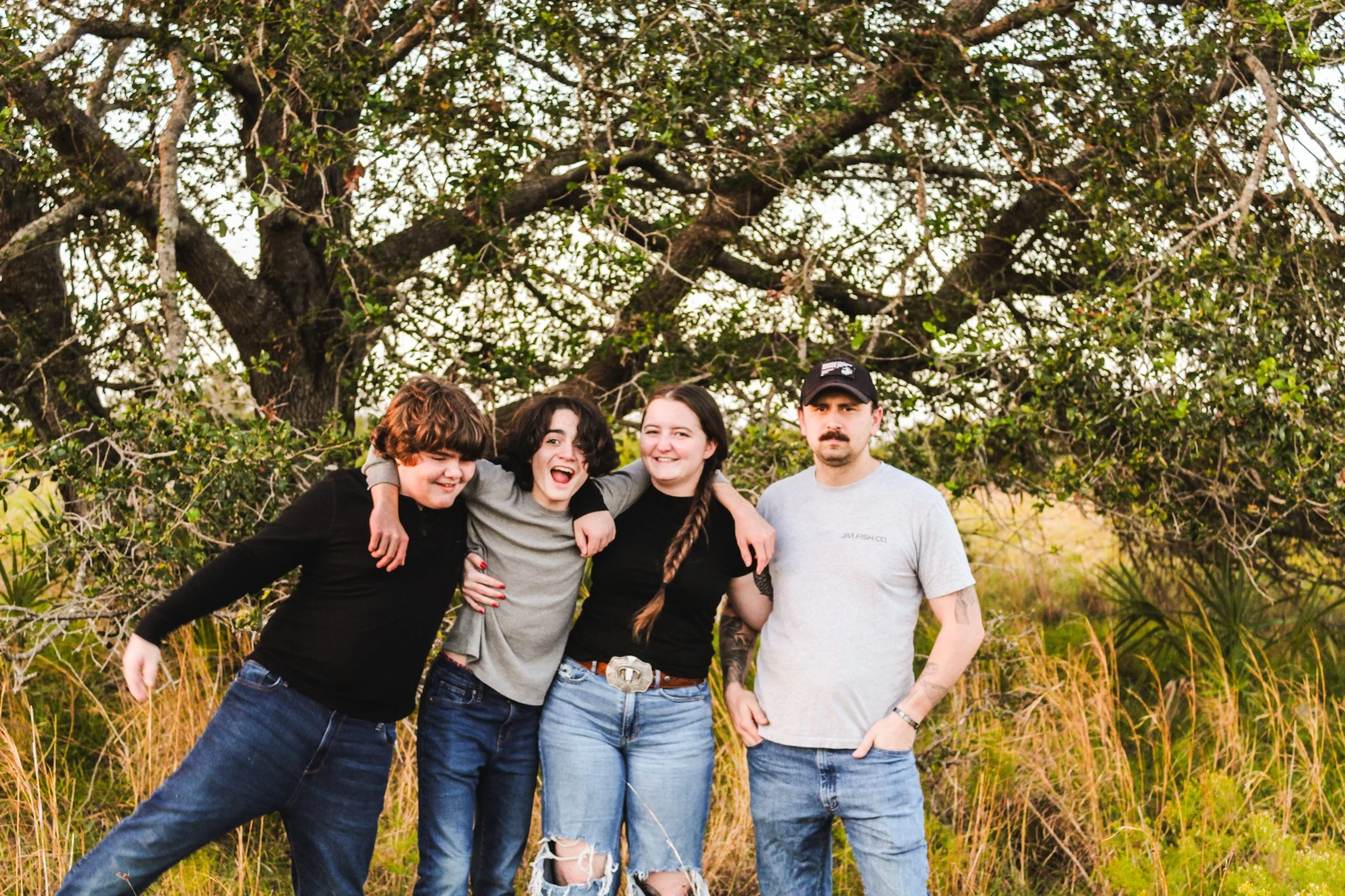 Four people standing outdoors in front of a large tree, smiling and embracing each other, with tall grass and bushes behind them.
