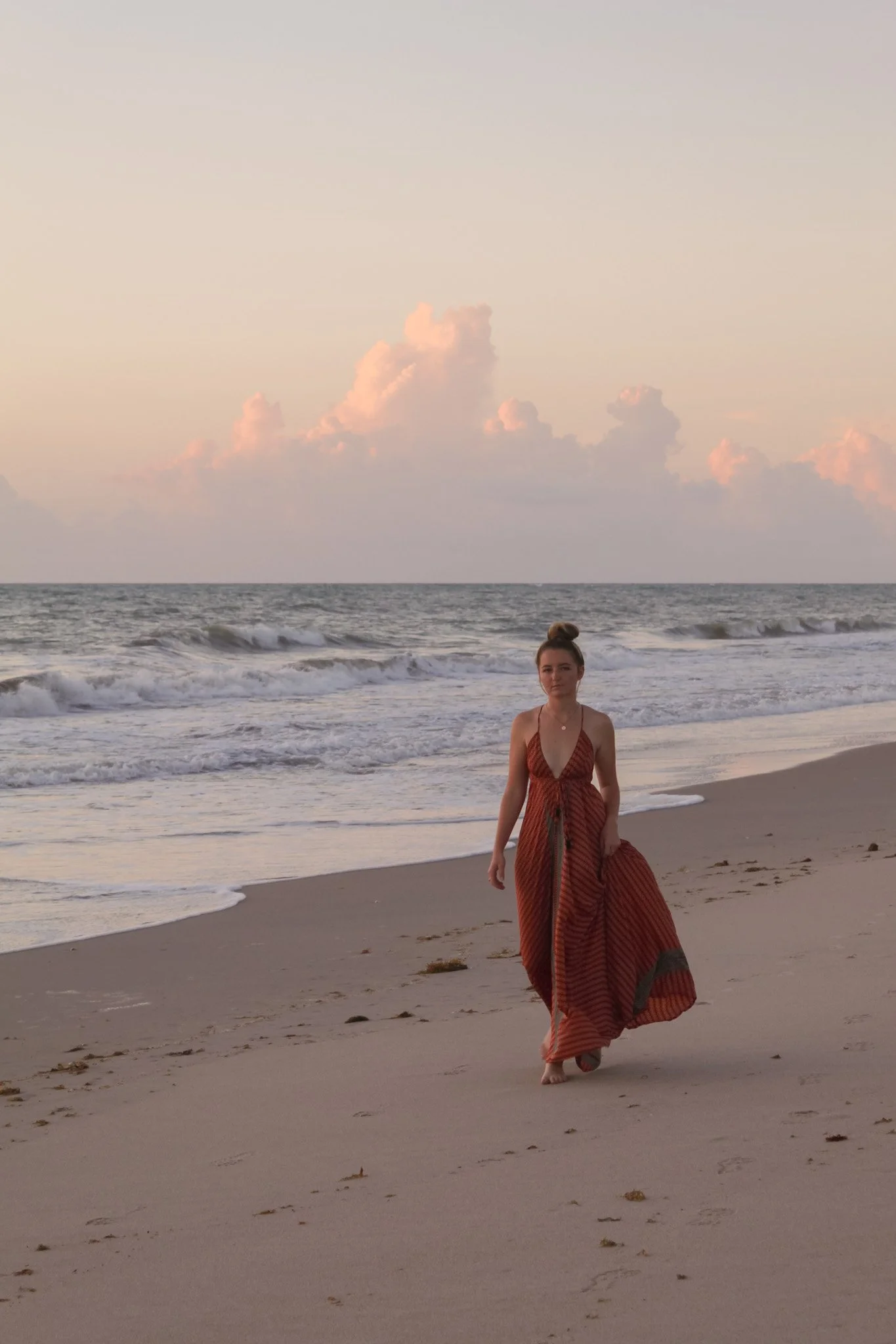 A woman in a brown and orange striped dress walking barefoot on a sandy beach at sunset with clouds in the sky and the ocean waves in the background.