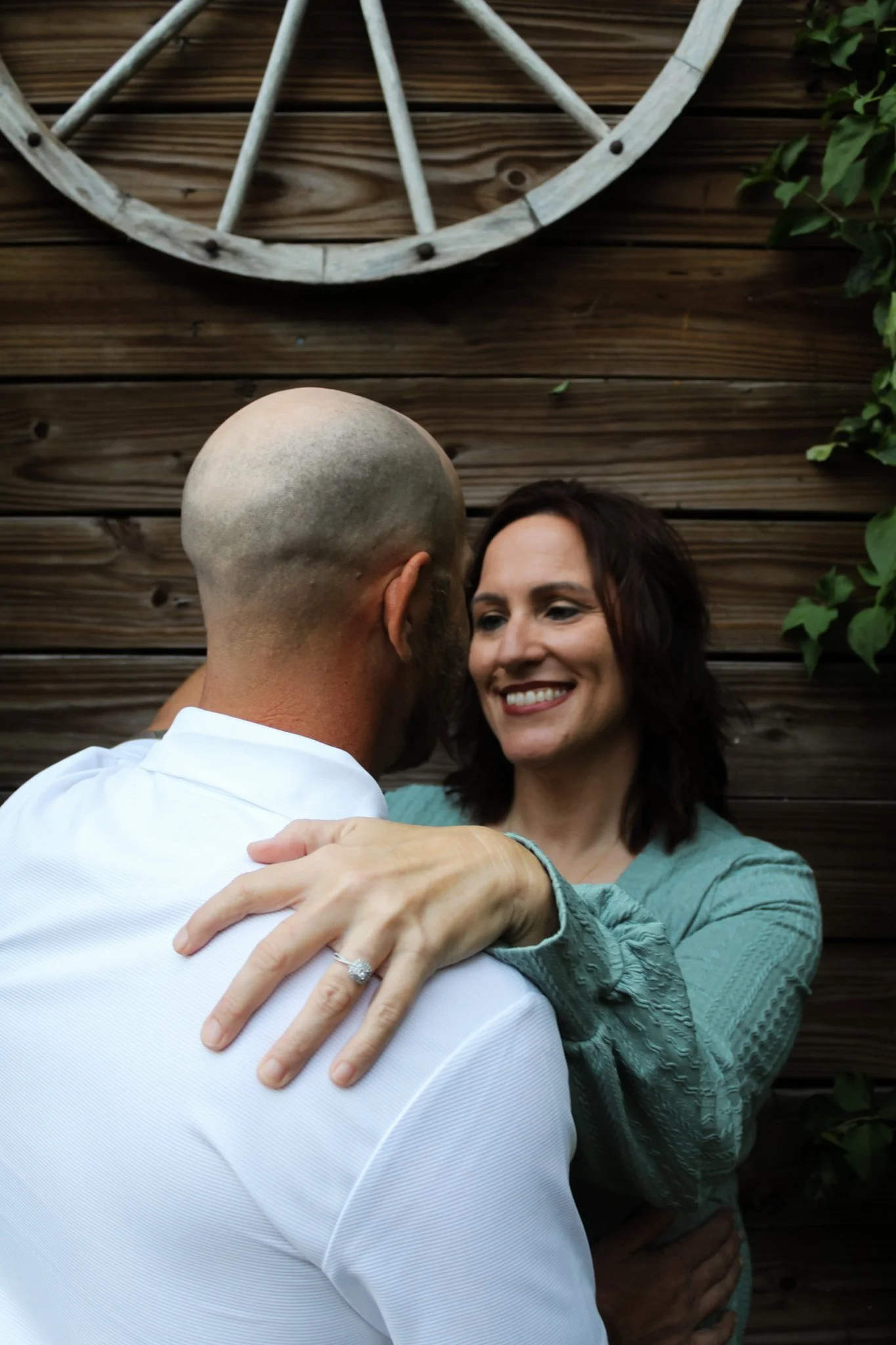 A woman with dark brown hair and a teal top embraces a bald man in a white shirt, smiling together against a wooden background with a decorative wagon wheel and green leafy plants.