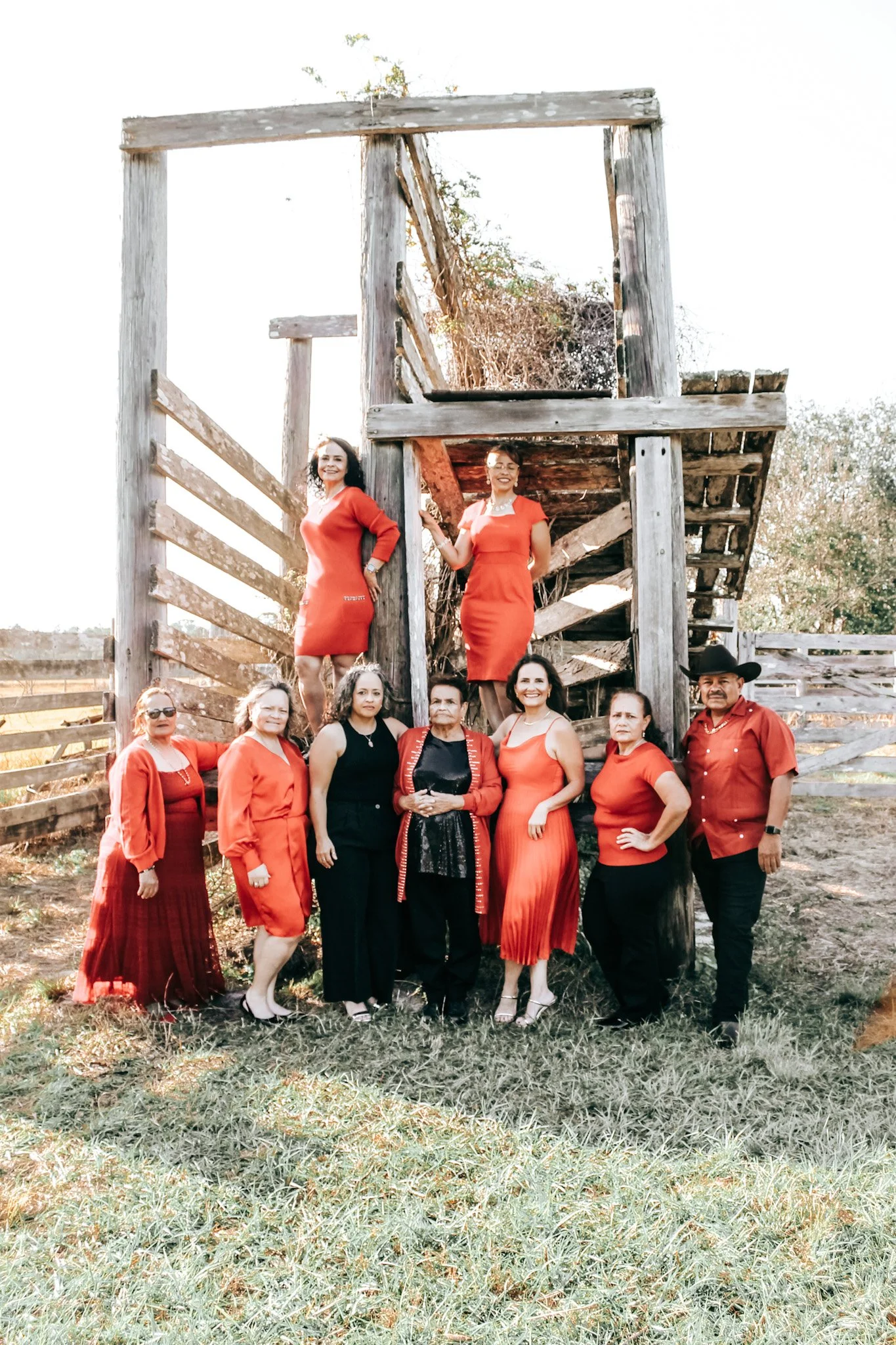 Group of nine people, mostly women, standing and sitting near an old wooden structure outdoors on grass, dressed in red and black attire, with some wearing dresses and others in shirts and pants.