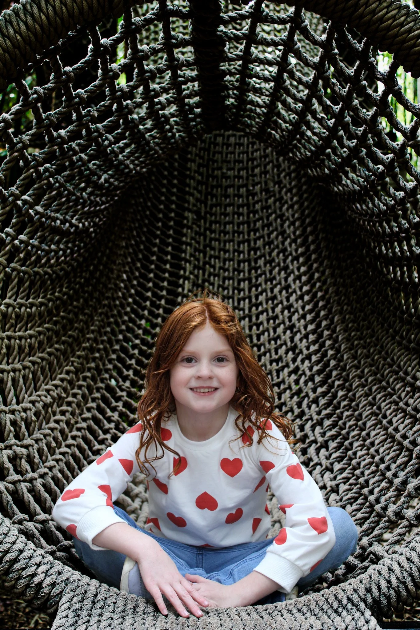 A young girl with curly red hair and a white long-sleeve shirt with red hearts, sitting cross-legged inside a large woven rope hammock in a natural outdoor setting.