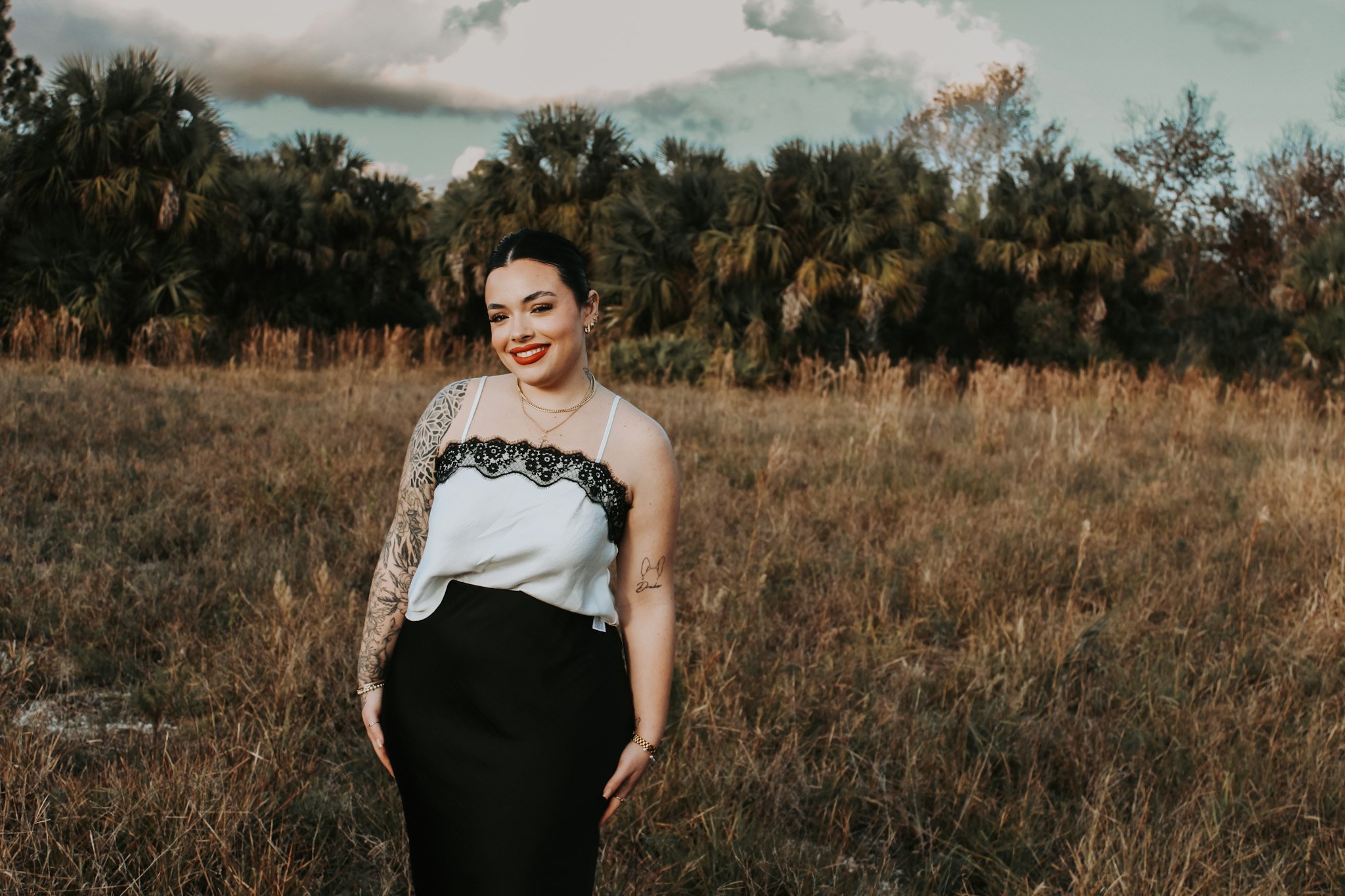 A woman with dark hair and tattoos standing in a grassy field with trees in the background during sunset.