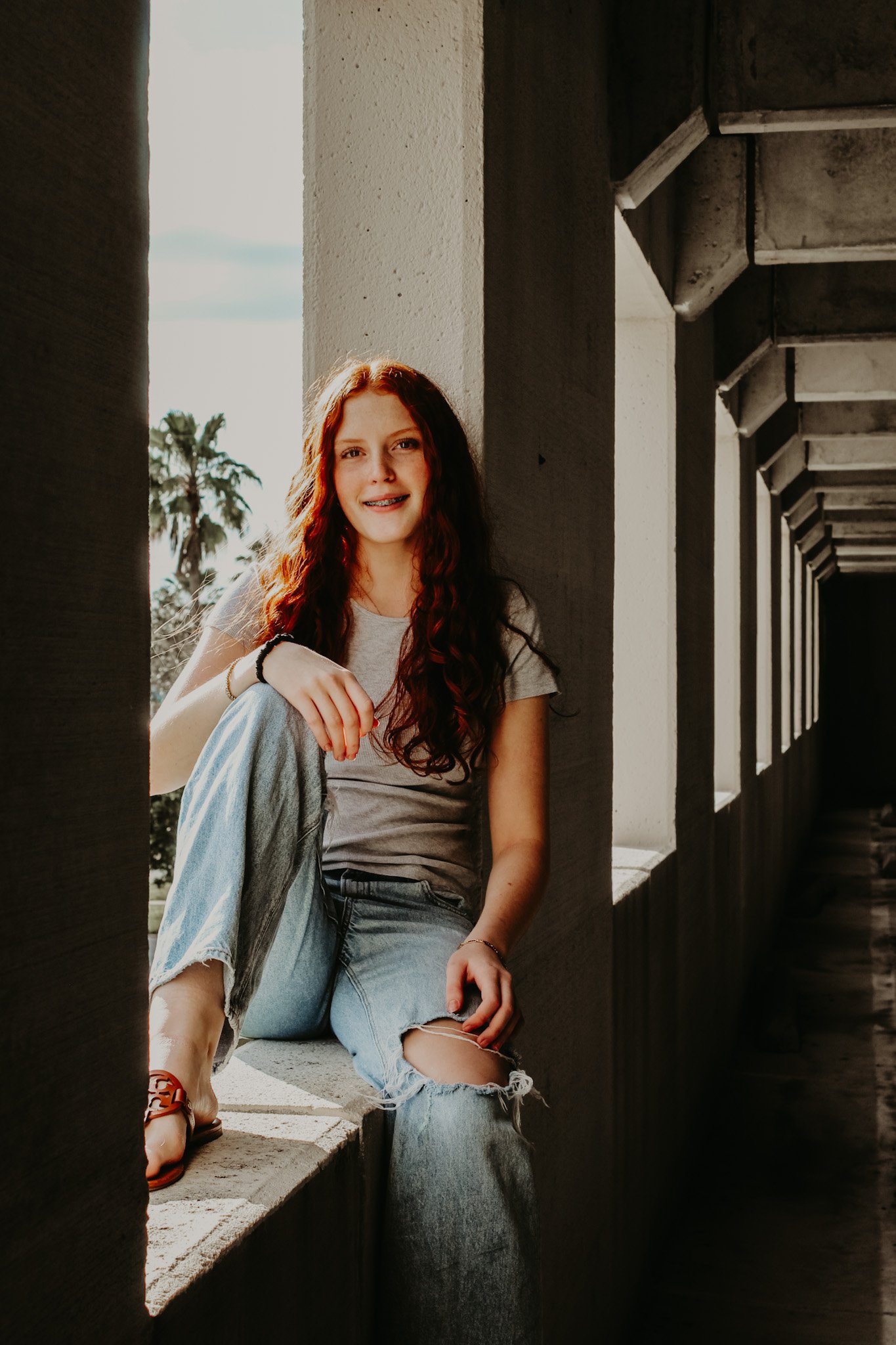 Young woman with long dark hair sitting on a ledge in a concrete structure, sunlight on her face, wearing a gray T-shirt and ripped jeans, smiling at the camera.