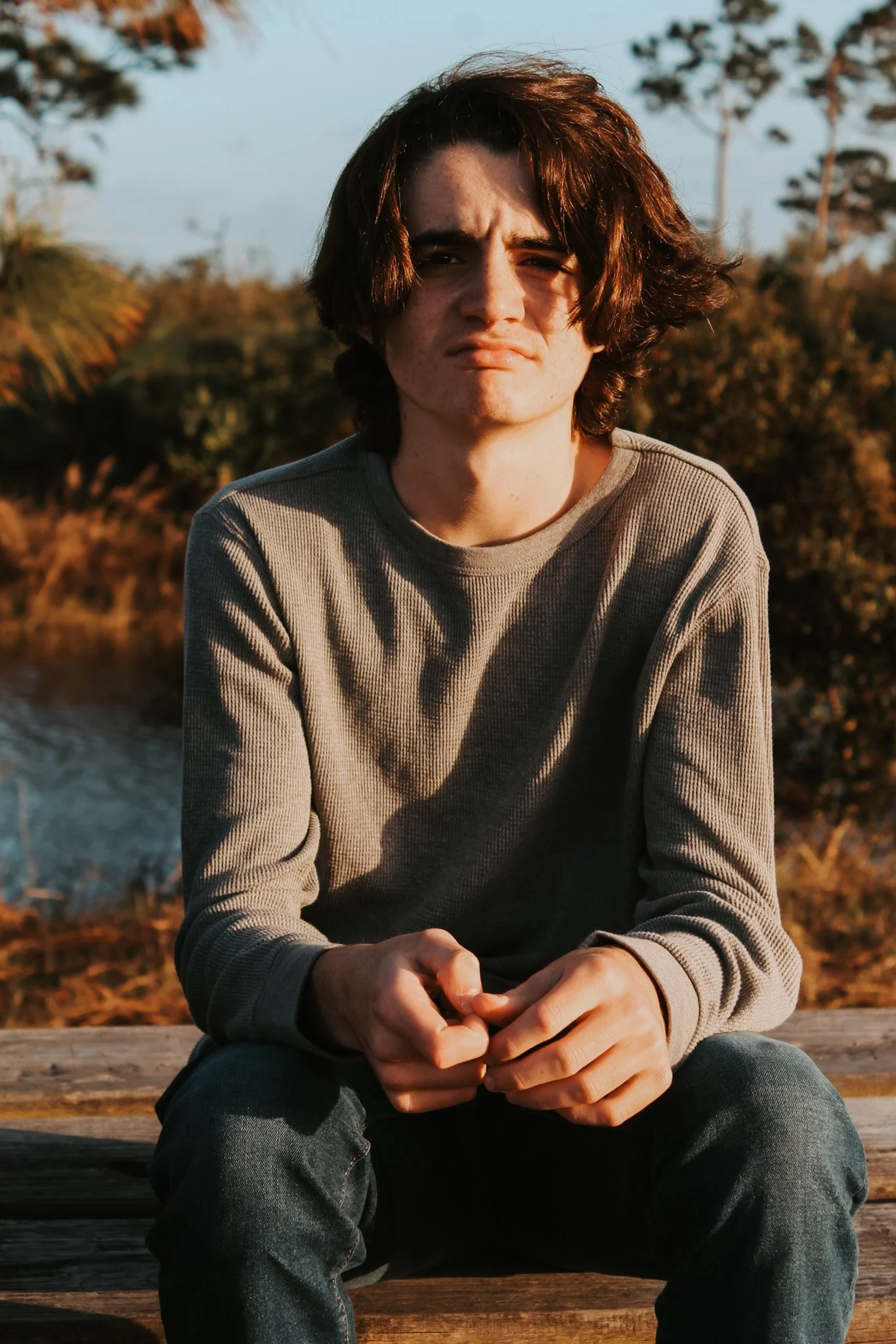 A young man with dark, wavy hair sitting on a wooden bench outdoors during sunset, wearing a gray long-sleeve shirt and jeans, with trees and water in the background.