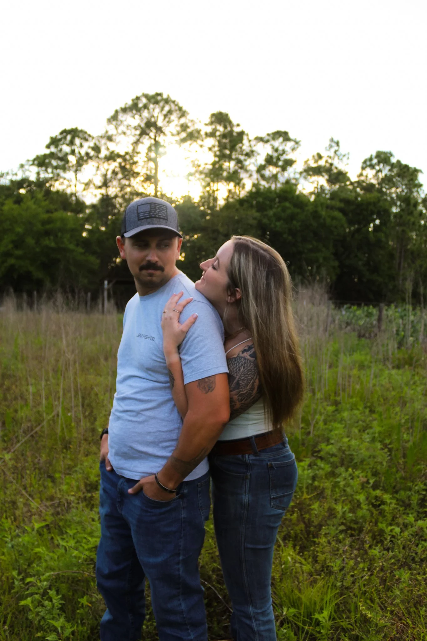 A couple standing in a grassy field during sunset, with trees in the background. The woman is leaning on the man's shoulder, and the man is looking off into the distance.