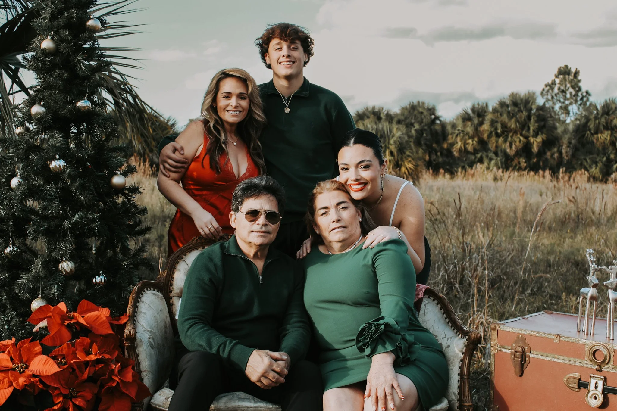 A family of five posing outdoors during Christmas, with a decorated Christmas tree and poinsettias nearby. They are dressed in festive clothing, sitting and standing on a vintage couch in a field with palm trees in the background.