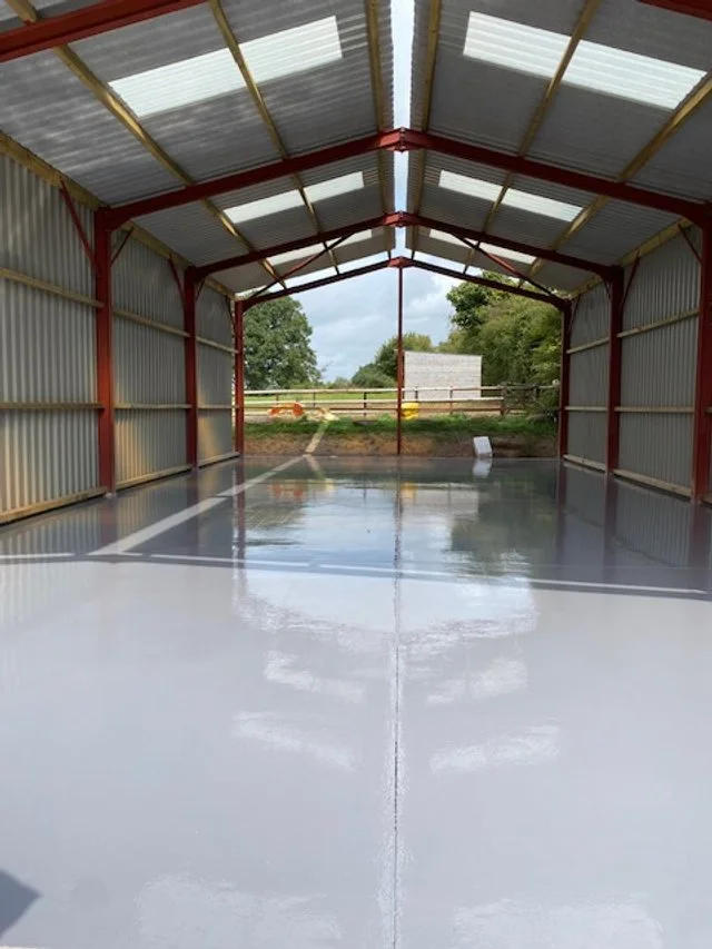 Inside of a newly painted, empty metal farm building or covered structure with a shiny floor and open sides, overlooking a grassy area with trees and a small building in the background.Resin floors The Wye Valley, Herefordshire, South Wales
