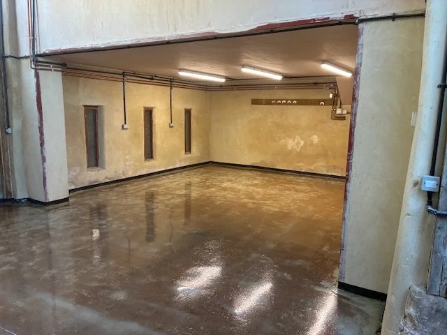 Empty indoor space with polished concrete floor, yellow walls, four narrow vertical windows, ceiling-mounted fluorescent lights, and exposed electrical wiring and fixtures.