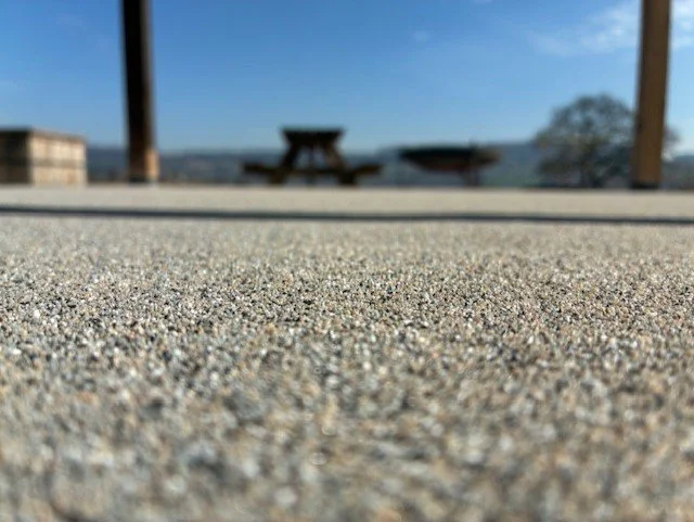 Close-up of a textured concrete surface with a blurred background featuring wooden tables and a scenic landscape under a blue sky. Resin floors The Wye Valley, Herefordshire, South Wales, West Midlands, west, cardiff, birmingham, bristol
