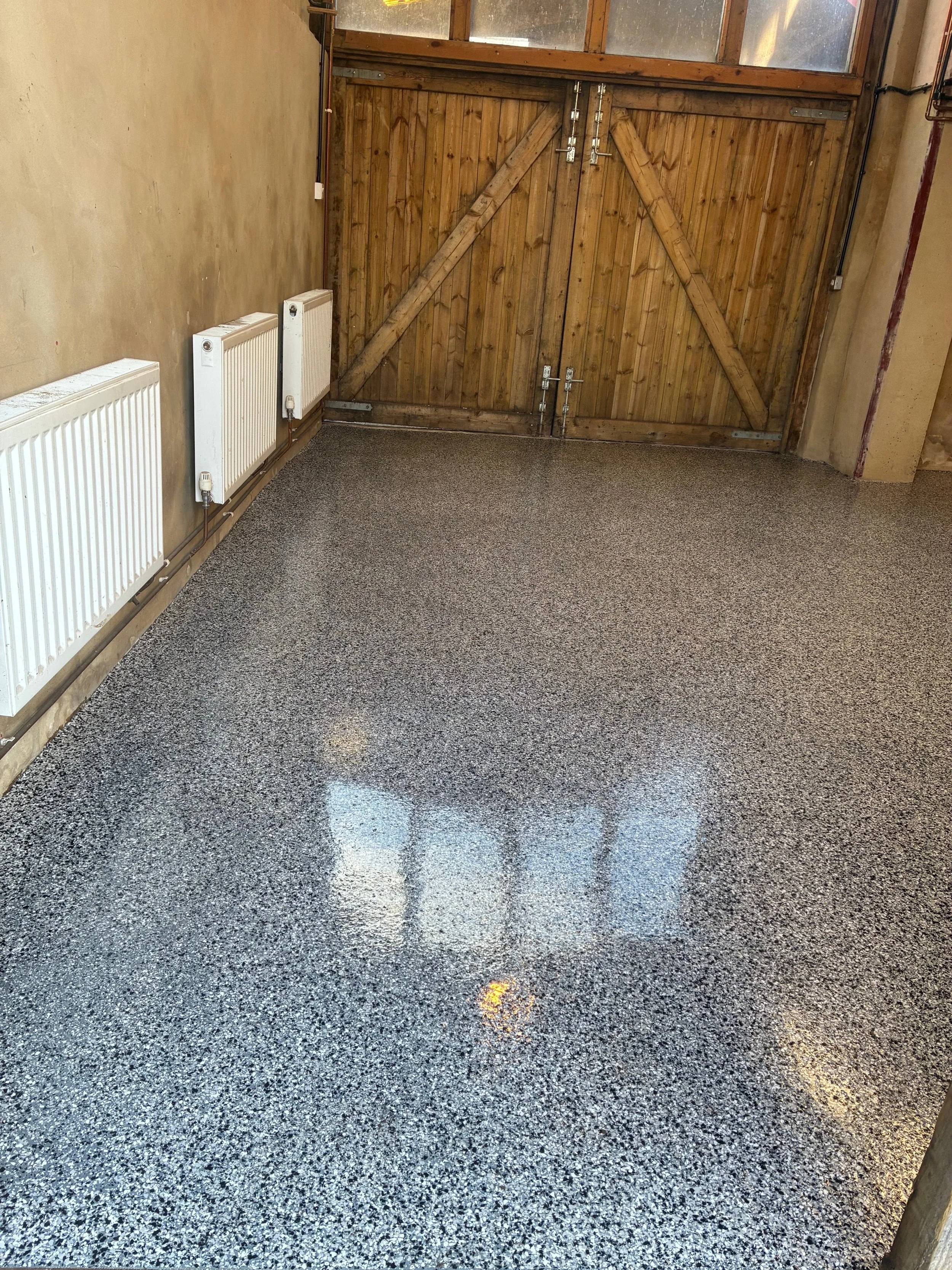 Empty garage with speckled epoxy floor, wooden garage door, and white radiators mounted on the beige wall.Resin floors The Wye Valley, Herefordshire, South Wales, West Midlands, west, cardiff, birmingham, bristol