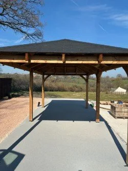 Open wooden pavilion with a shingled roof, supported by wooden pillars, on a concrete slab, with a clear blue sky and trees in the background.Resin floors The Wye Valley, Herefordshire, South Wales, West Midlands, west, cardiff, birmingham, bristol