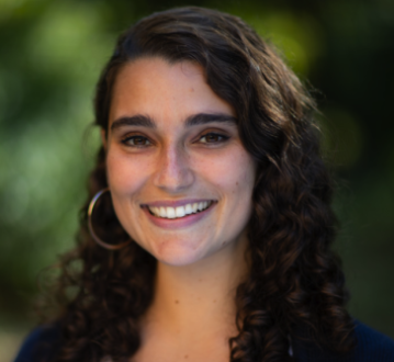 A young woman with curly brown hair smiling outdoors with a blurred green background.