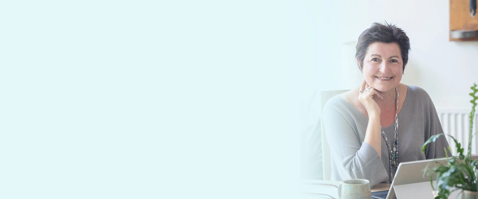 A smiling woman with short dark hair sitting at a table with a laptop, a cup, and some plants in a bright, modern room.