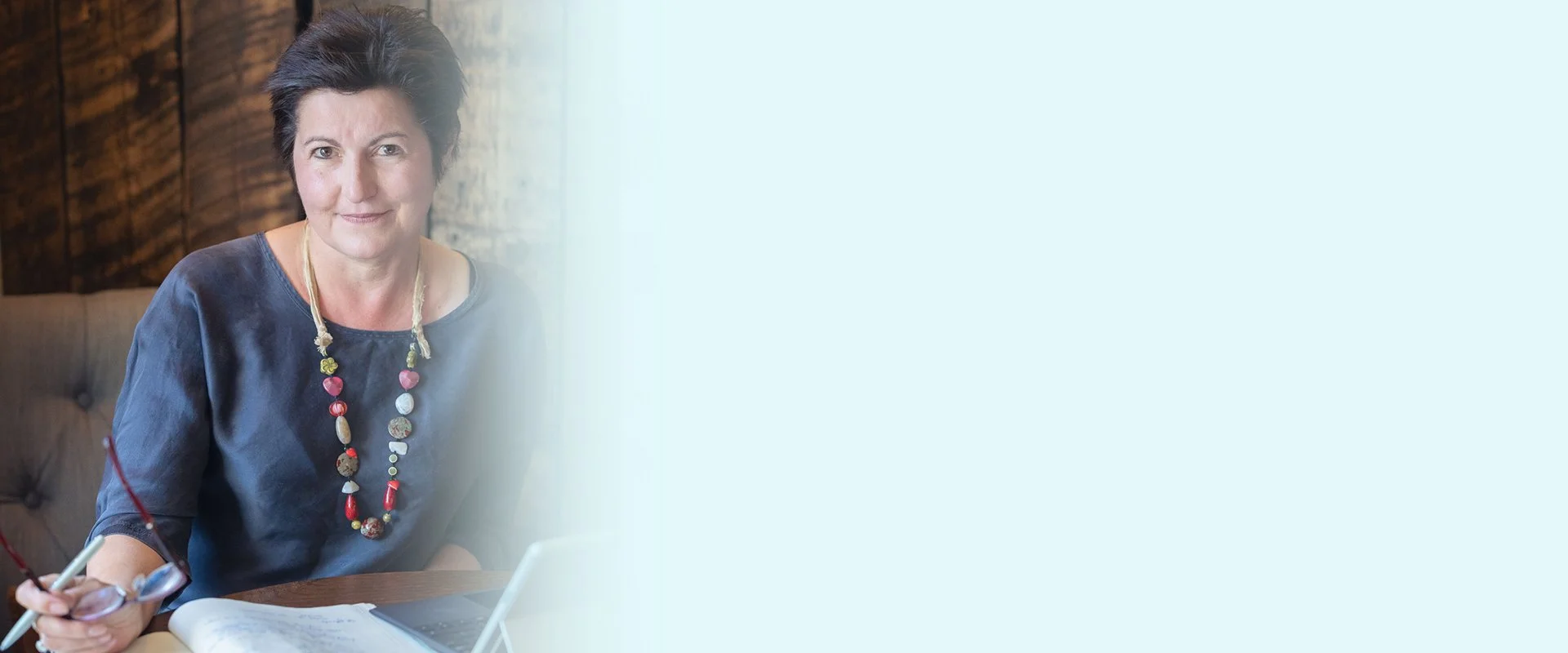 A woman with short dark hair wearing a dark blue shirt and a multi-colored beaded necklace sitting at a desk with a notepad, pen, and a glass of red wine.