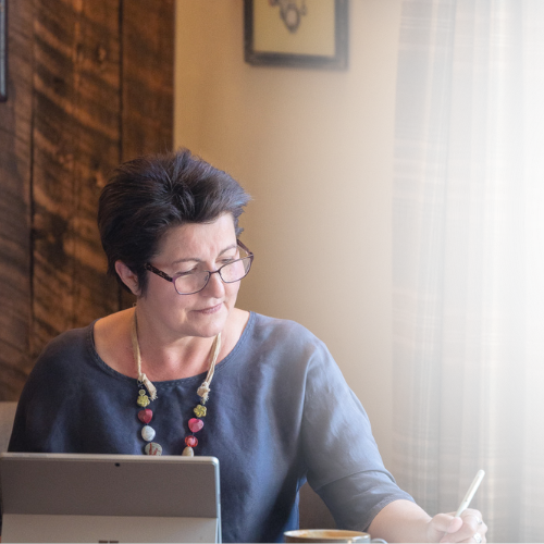 A woman with short dark hair and glasses working at a desk with a tablet, a pen, and a cup, sitting by a window with curtains.