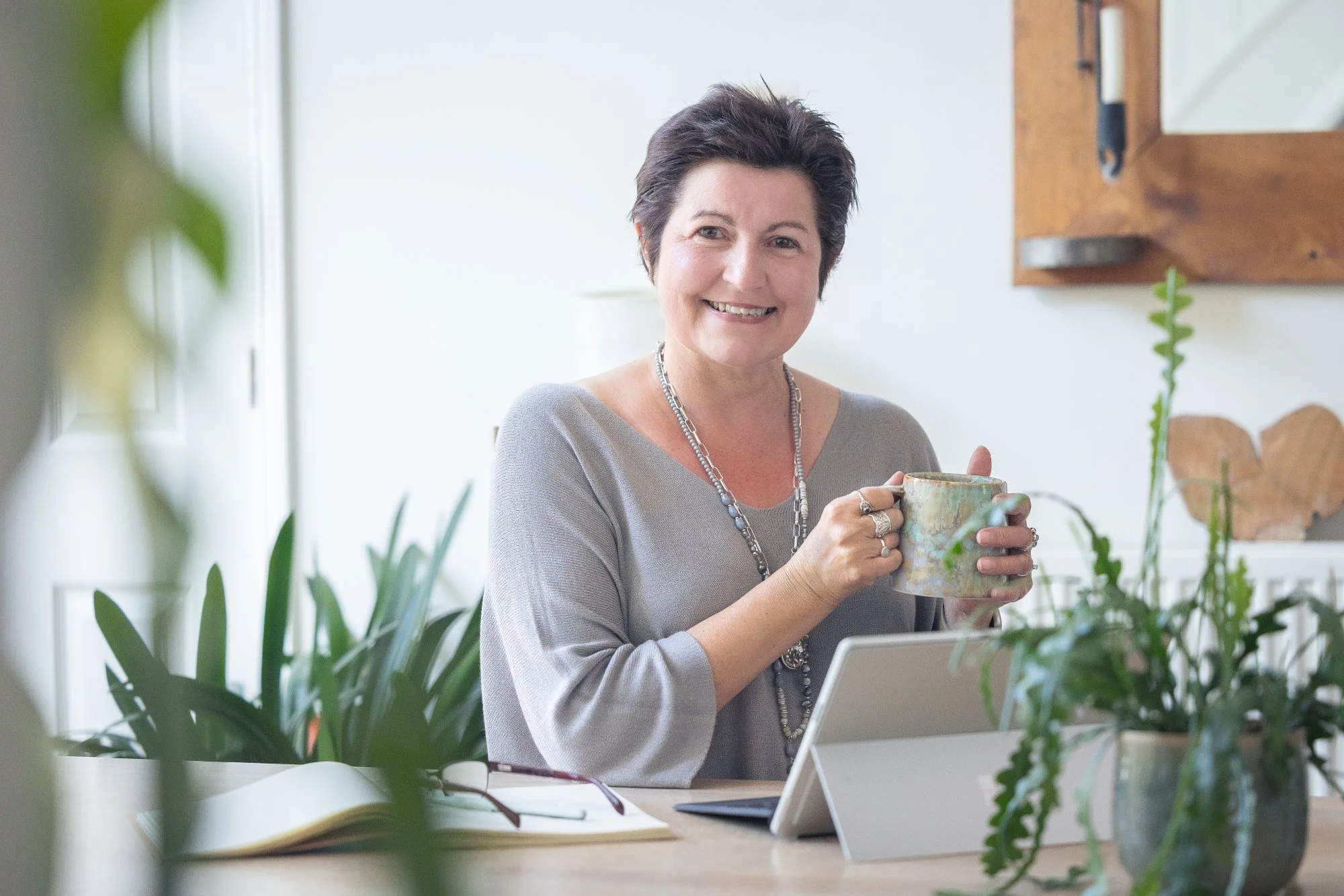 A smiling woman with short dark hair sitting at a desk holding a mug, surrounded by plants and a tablet.