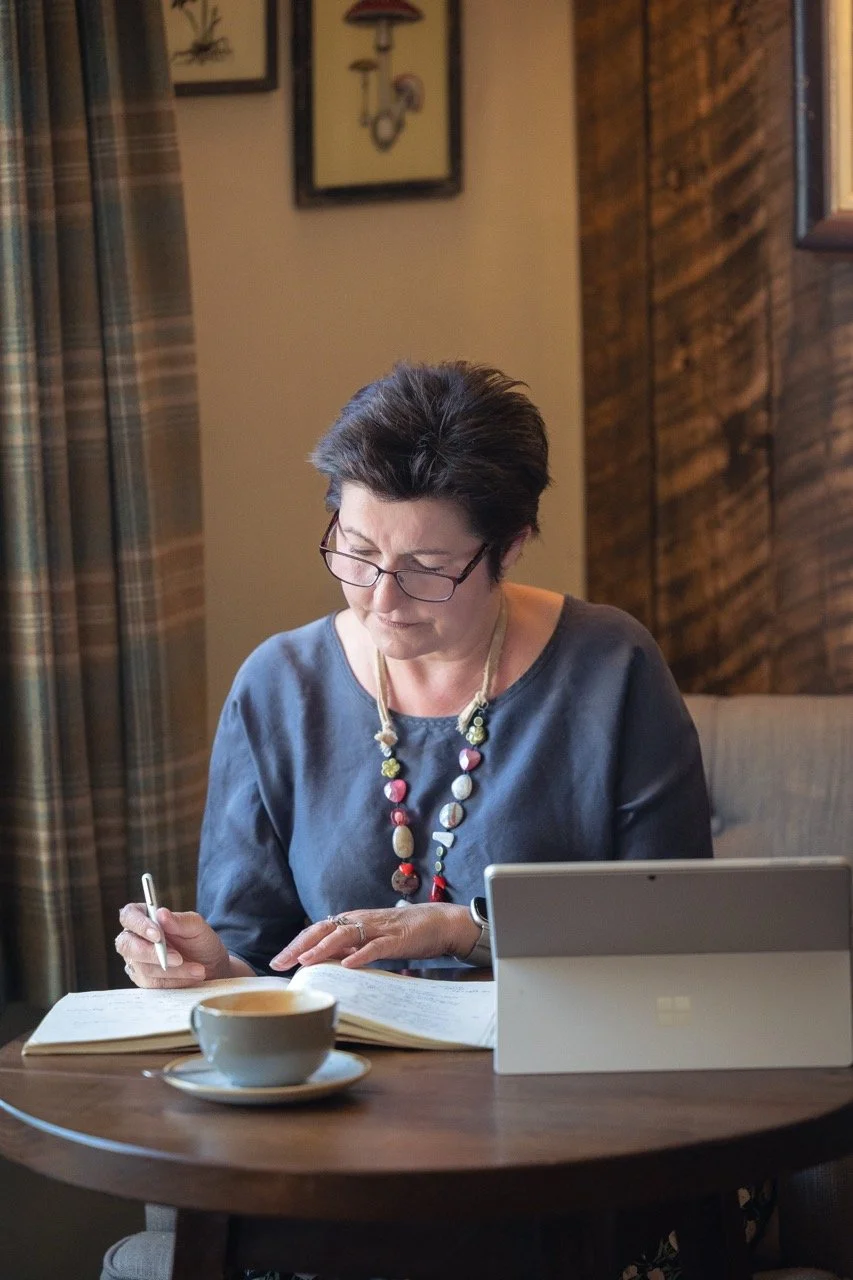 A woman with short dark hair and glasses sitting at a table, looking at a notebook, with a cup of coffee, a tablet, and a pen in her hands.