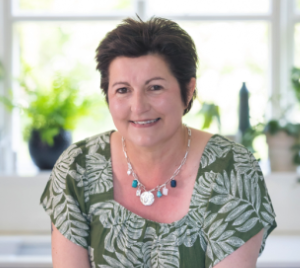 A smiling woman with short dark hair wearing a green patterned blouse and a colorful necklace, sitting indoors with a window and plants in the background.