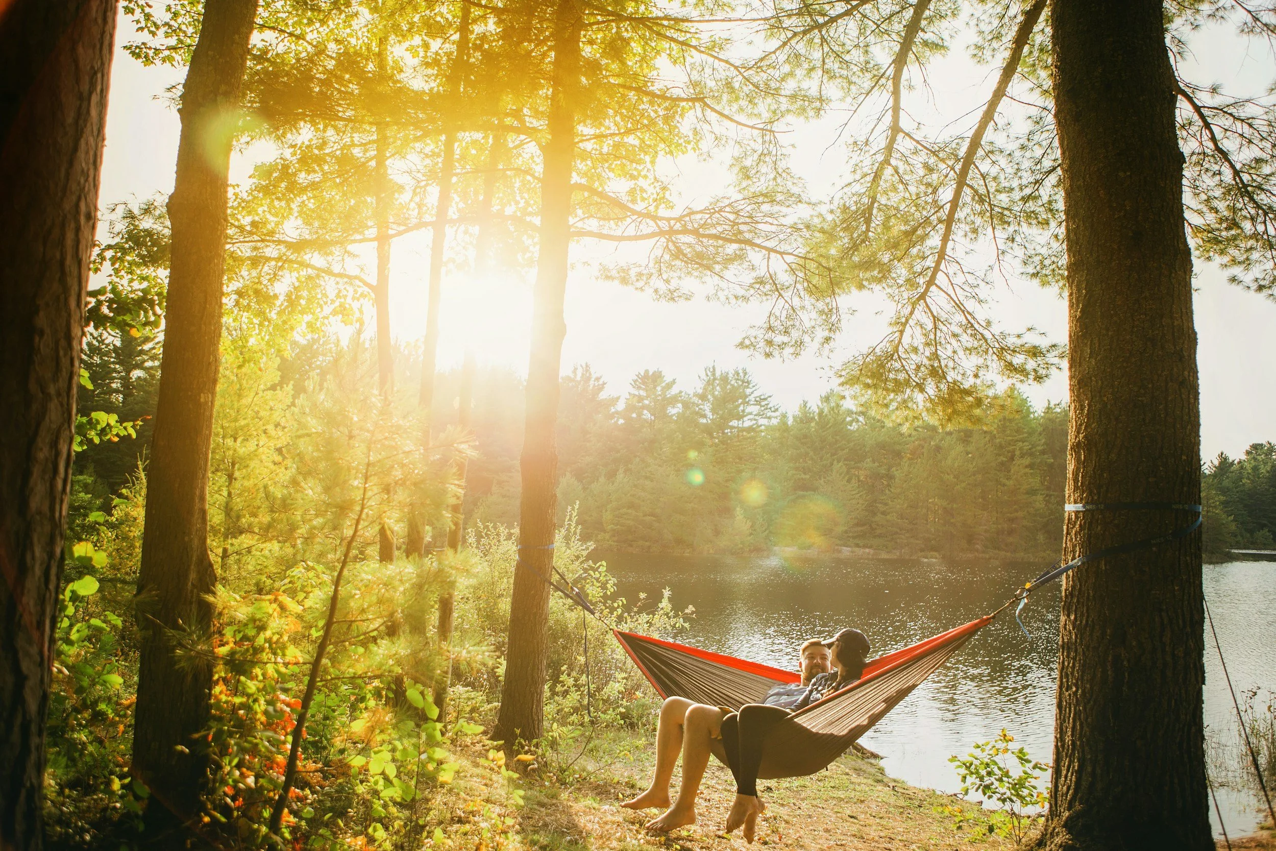 Two people relaxing in a hammock by a lake, surrounded by trees with sunlight filtering through.