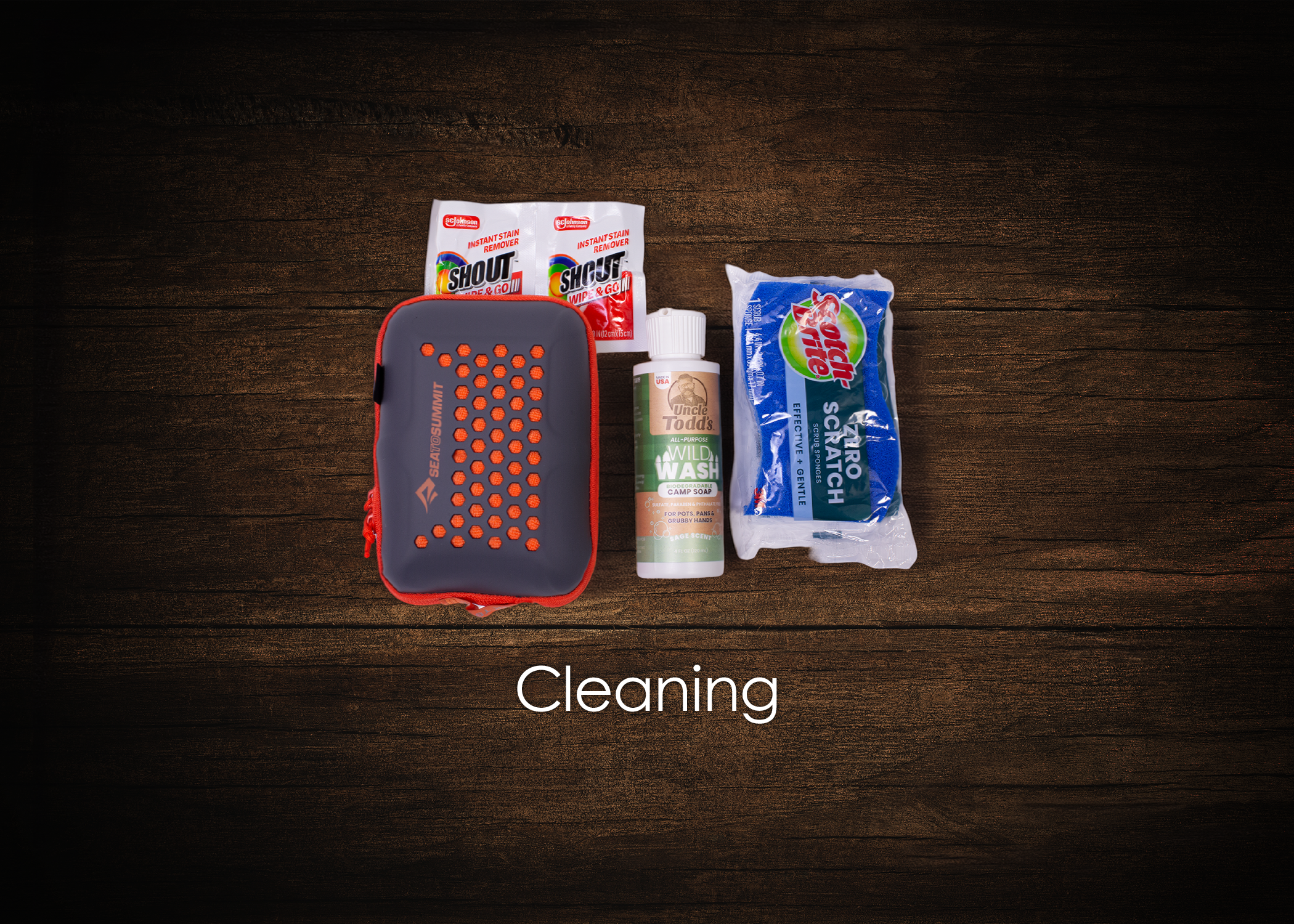 Various cleaning supplies on a wooden surface, including stain remover packs, a bottle of soap, a scrub sponge, and a carrying case.