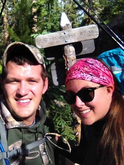A man and woman smiling outdoors, wearing backpacks and sunglasses, near a trail sign.