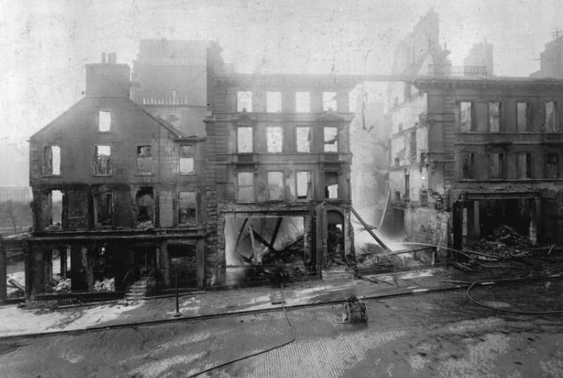 A black and white photo of a building with burned and damaged walls and windows in 1892, debris on the ground, and smoke or soot covering the scene.
