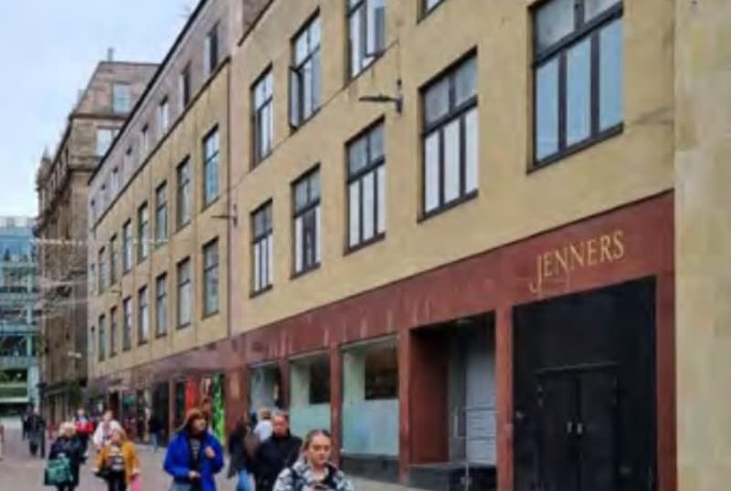 A city street with several pedestrians walking along the sidewalk. The building on the right side has a tan exterior with black-framed windows and a sign that reads 'JENNERS'.