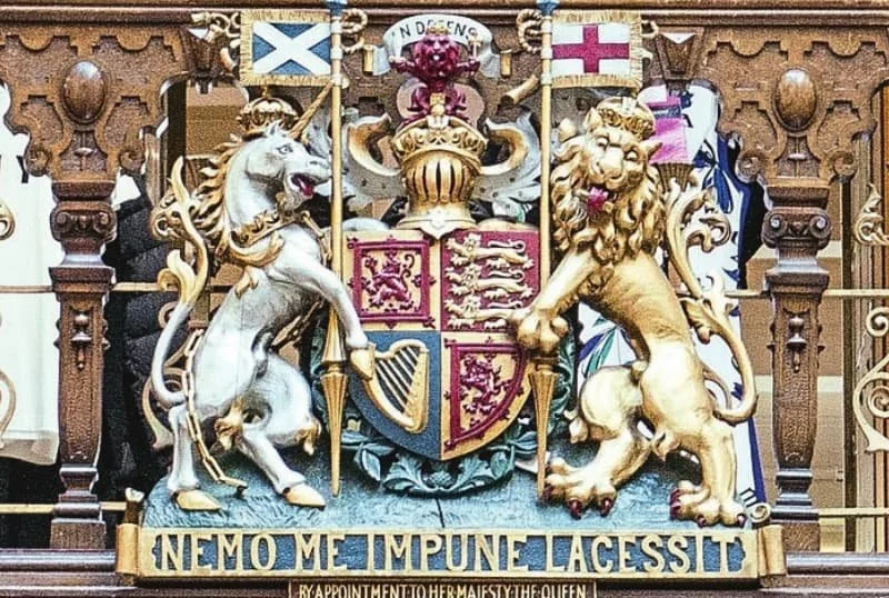The royal coat of arms of the United Kingdom featuring a lion and a unicorn, with a motto 'Nemo Me Impune Lacessit' at the bottom awarded as a Royal Warrant to the Jenners.
