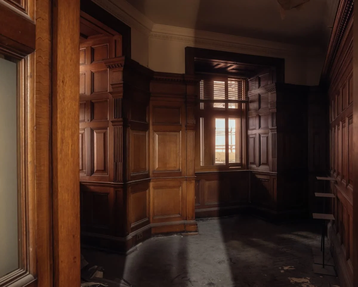 A room with dark wooden paneling and built-in bookshelves, sunlight coming through a large window, and a worn carpeted floor.