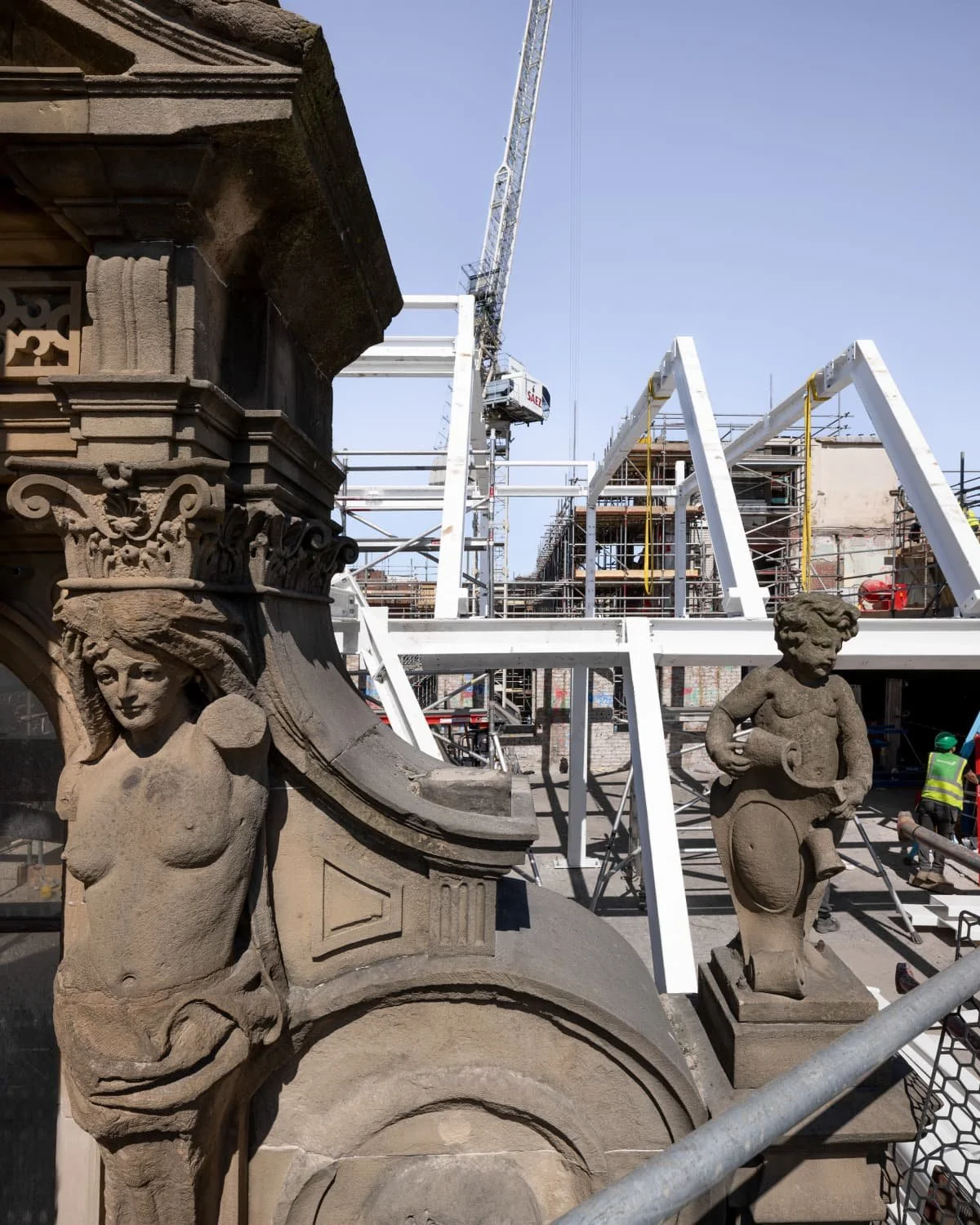 Close-up of ornate stone sculptures and carvings on a historical building under restoration, with scaffolding and a construction crane in the background.