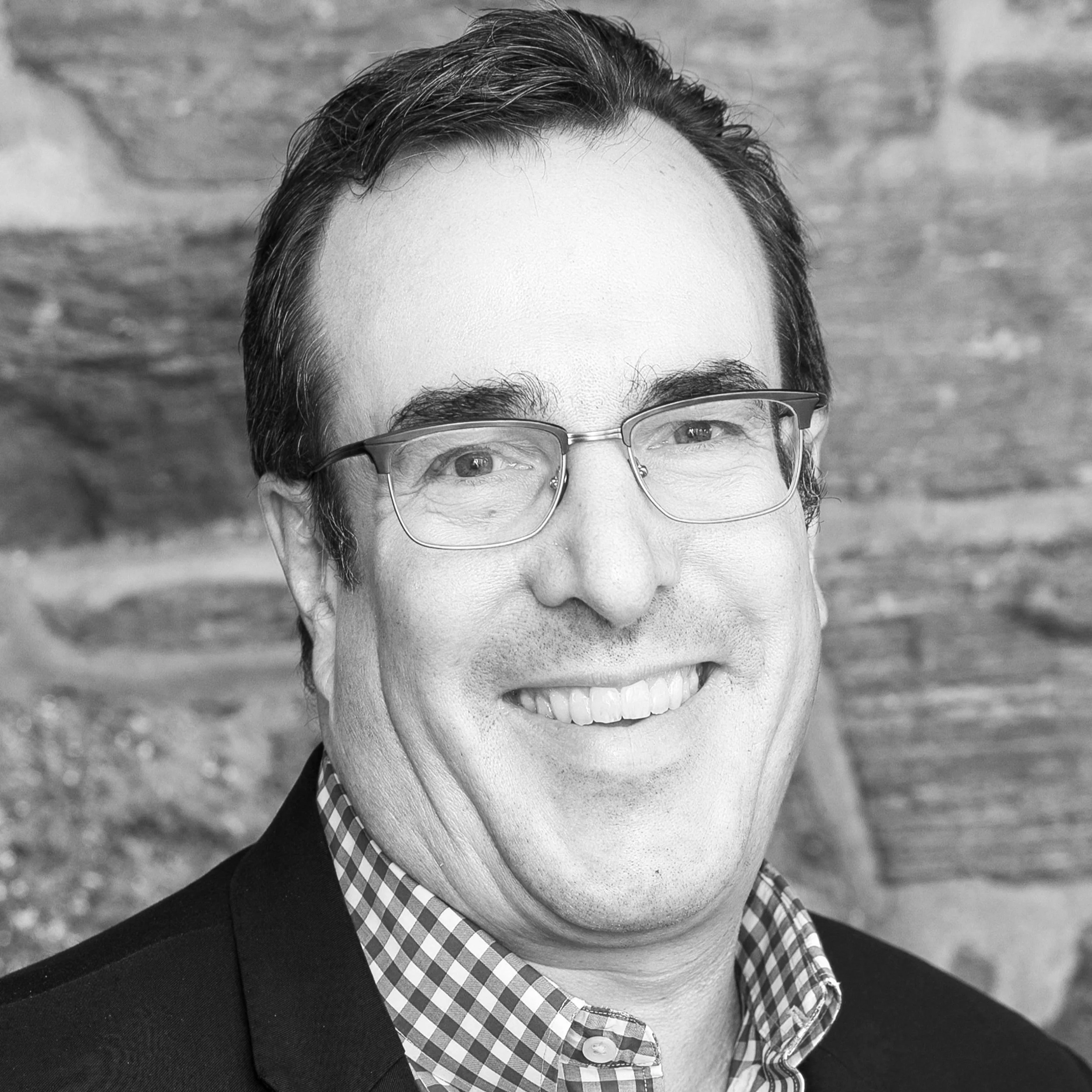A black-and-white close-up portrait of a smiling man with glasses, dressed in a checkered shirt and a dark blazer, with a textured brick wall background.